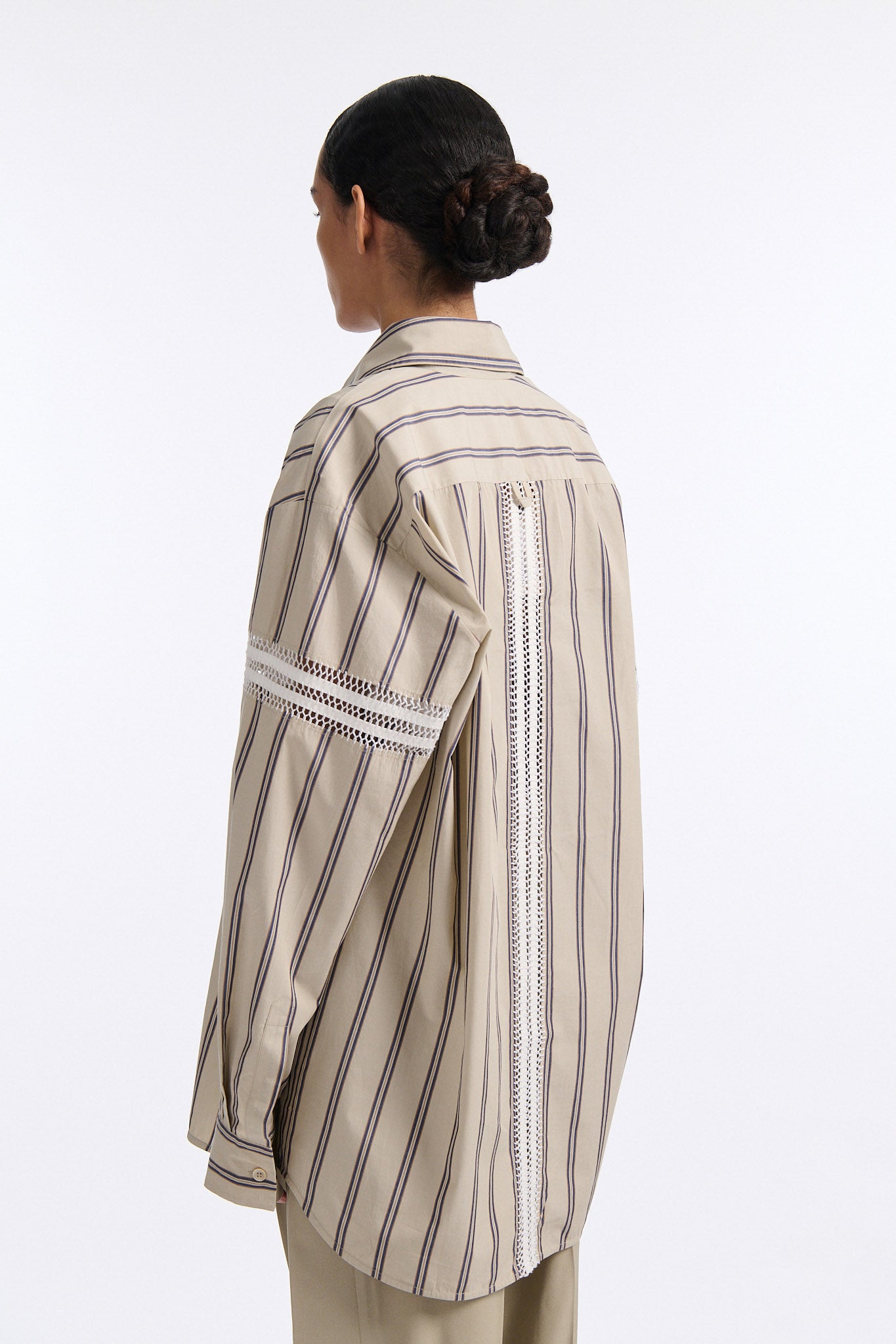 Female model with slicked back black hair in a bun wears vertical striped ecru button-down shirt with hand stitched stripes around either upper-arm. Model also wears ecru pleated trouser with extra panel on sides for volume.