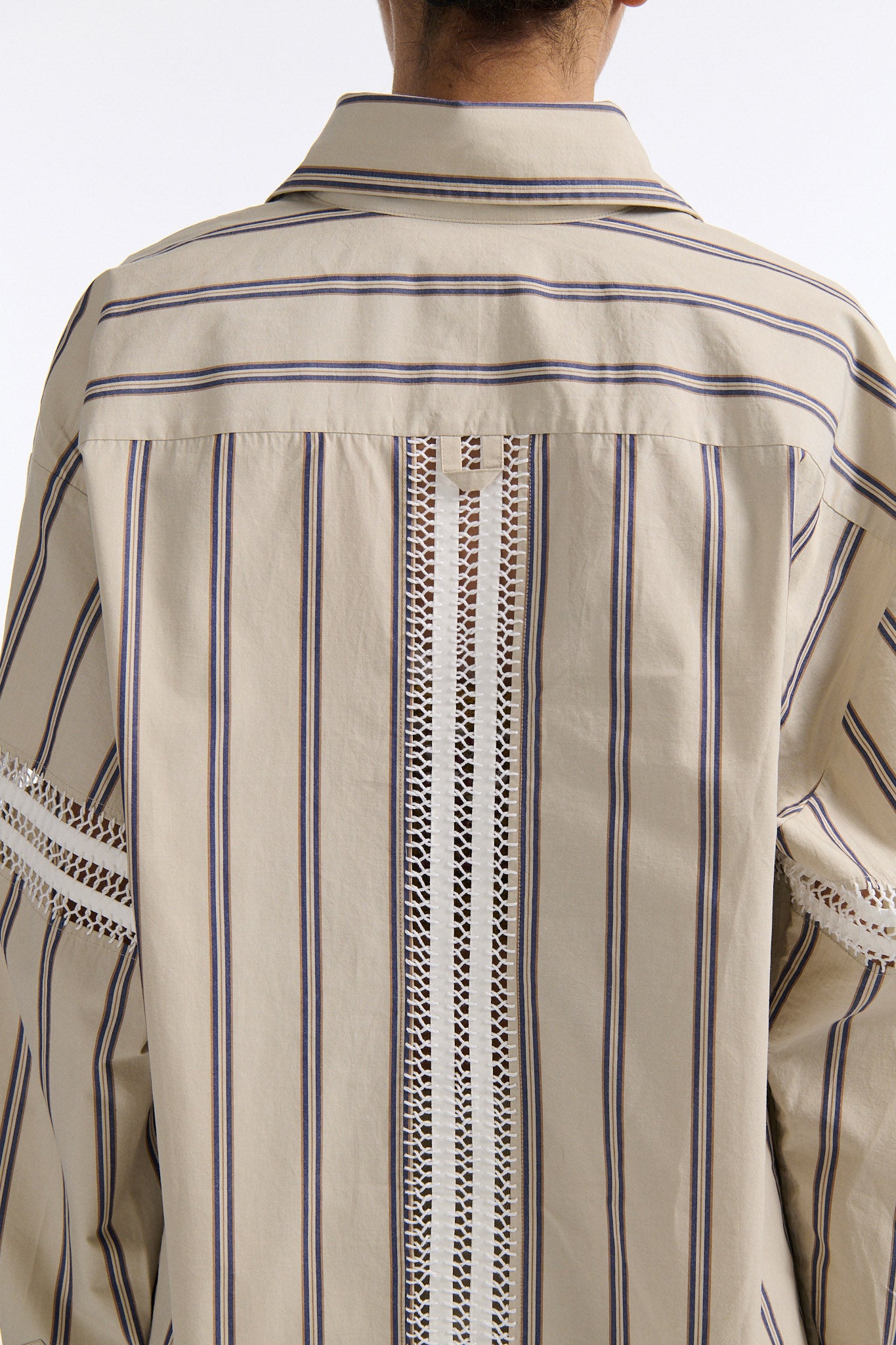 Female model with slicked back black hair in a bun wears vertical striped ecru button-down shirt with hand stitched stripes around either upper-arm. Model also wears ecru pleated trouser with extra panel on sides for volume.