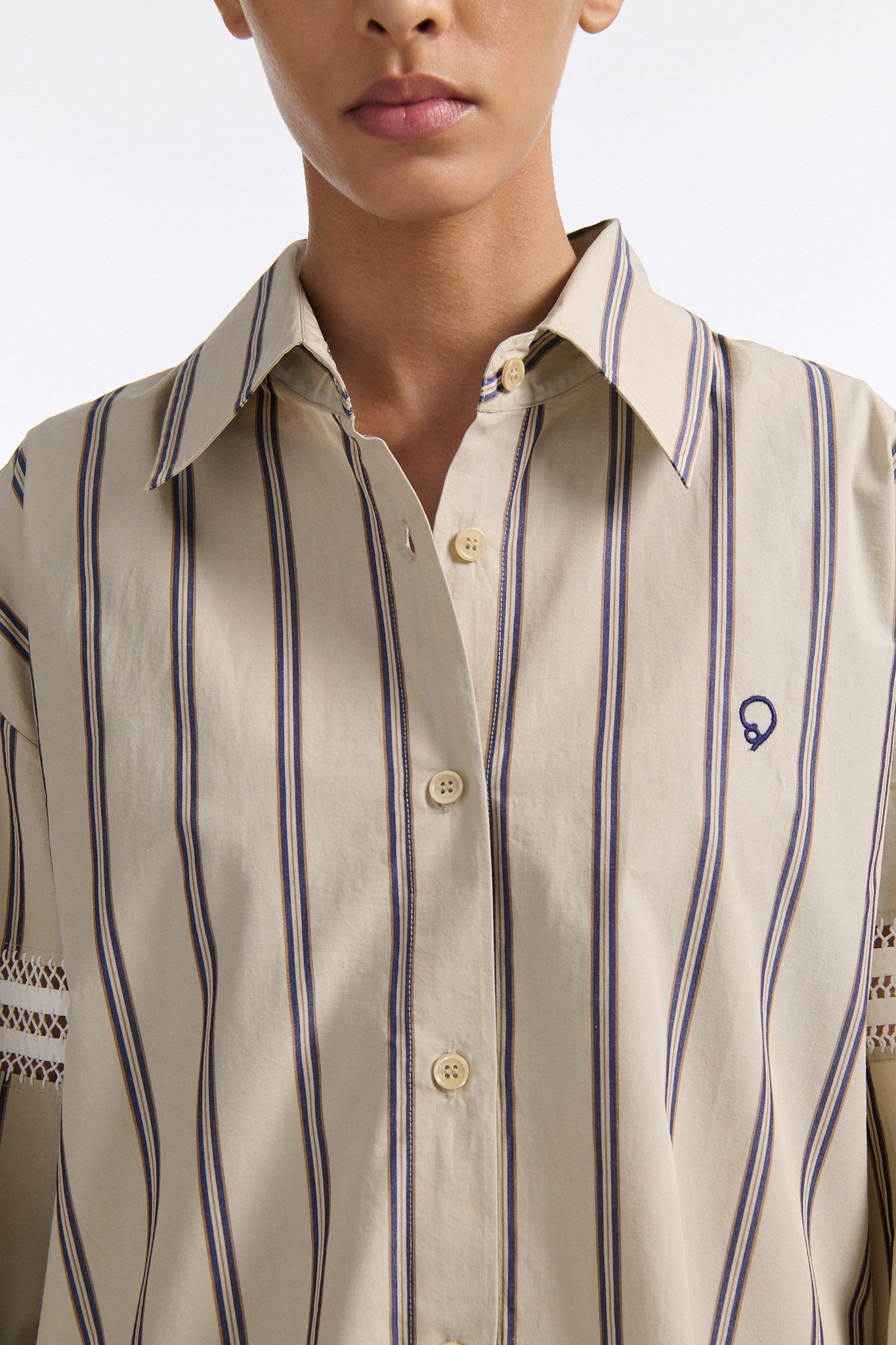 Female model with slicked back black hair in a bun wears vertical striped ecru button-down shirt with hand stitched stripes around either upper-arm. Model also wears ecru pleated trouser with extra panel on sides for volume.