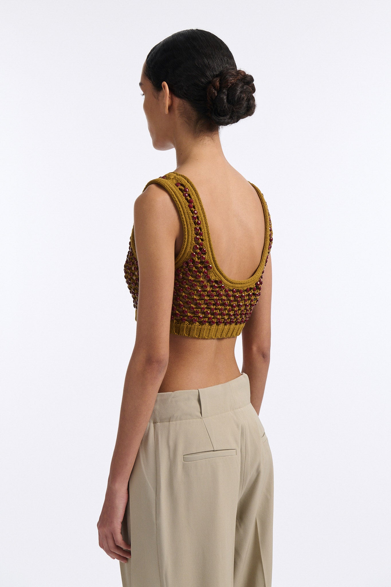 Female model with slicked back black hair in a bun wears cumin colored chunky mesh bralette with maroon hand-sewn crystals. Model also wears beige wide pleated trousers with additional panel at sides to increase volume.