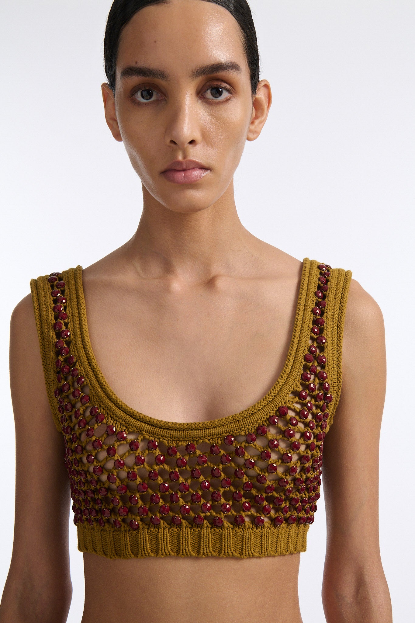 Female model with slicked back black hair in a bun wears cumin colored chunky mesh bralette with maroon hand-sewn crystals. Model also wears beige wide pleated trousers with additional panel at sides to increase volume.