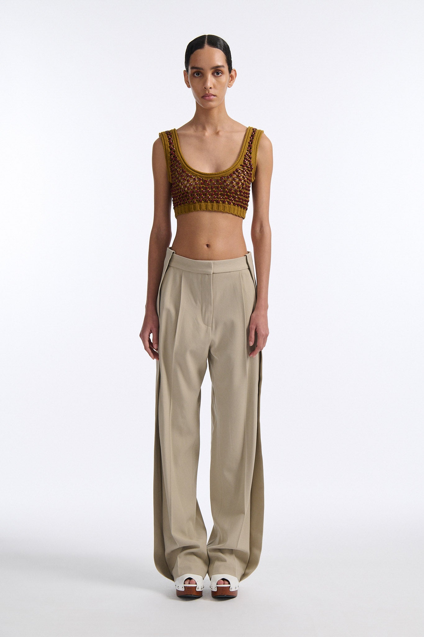 Female model with slicked back black hair in a bun wears cumin colored chunky mesh bralette with maroon hand-sewn crystals. Model also wears beige wide pleated trousers with additional panel at sides to increase volume.