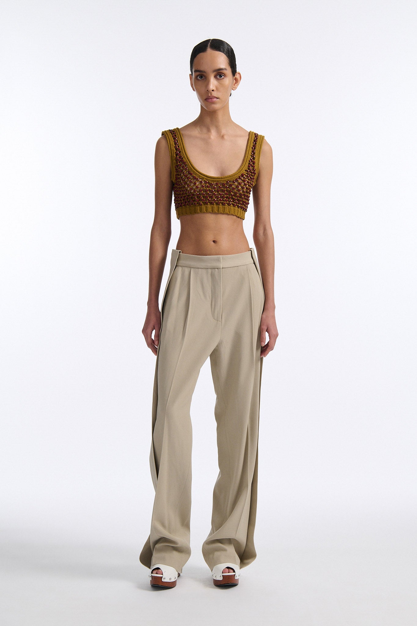 Female model with slicked back black hair in a bun wears cumin colored chunky mesh bralette with maroon hand-sewn crystals. Model also wears beige wide pleated trousers with additional panel at sides to increase volume.