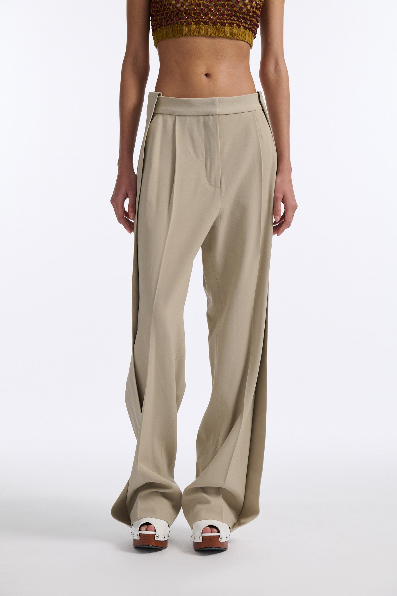 Female model with slicked back black hair in a bun wears cumin colored chunky mesh bralette with maroon hand-sewn crystals. Model also wears beige wide pleated trousers with additional panel at sides to increase volume.