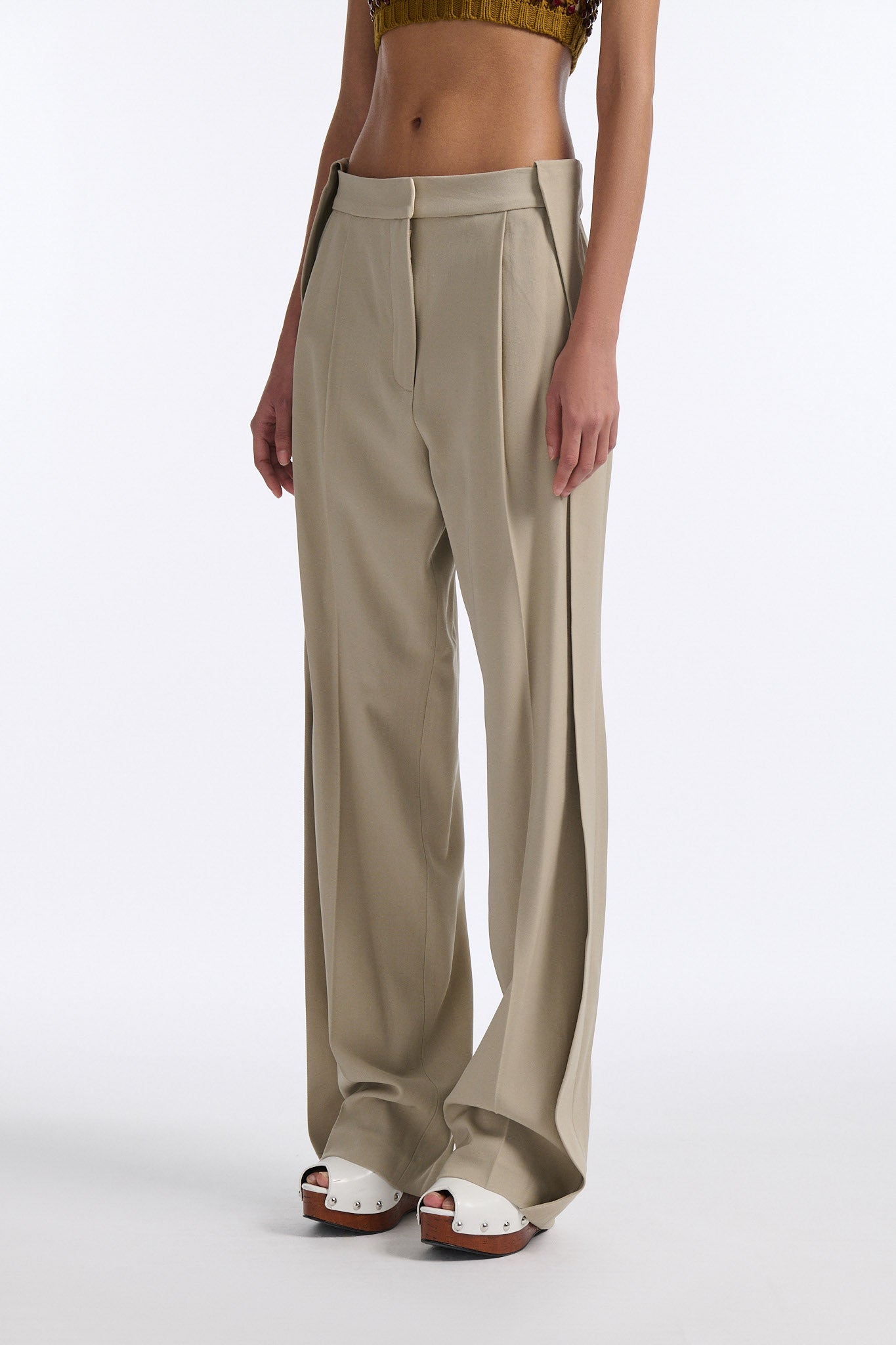 Female model with slicked back black hair in a bun wears cumin colored chunky mesh bralette with maroon hand-sewn crystals. Model also wears beige wide pleated trousers with additional panel at sides to increase volume.