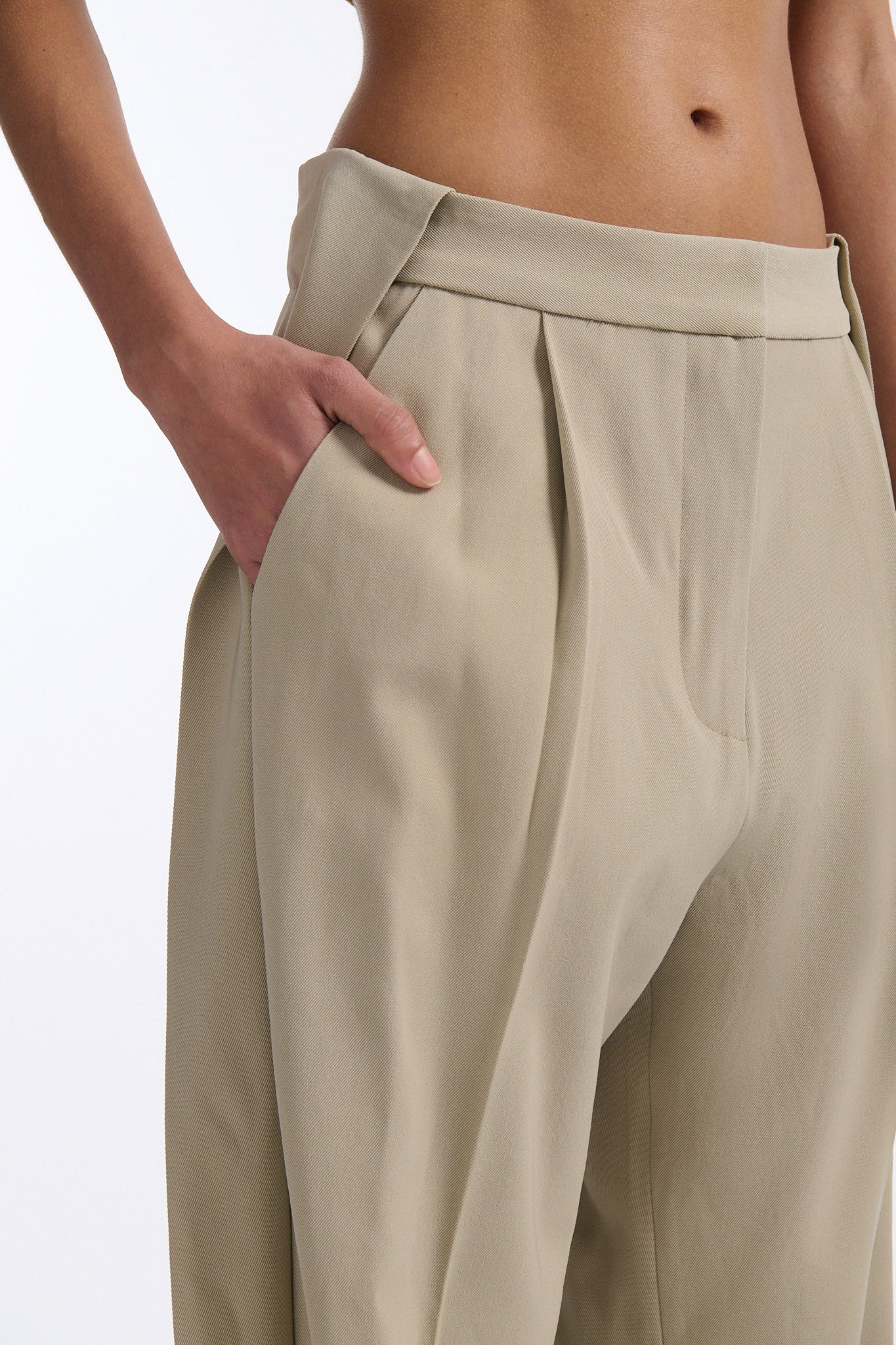 Female model with slicked back black hair in a bun wears cumin colored chunky mesh bralette with maroon hand-sewn crystals. Model also wears beige wide pleated trousers with additional panel at sides to increase volume.