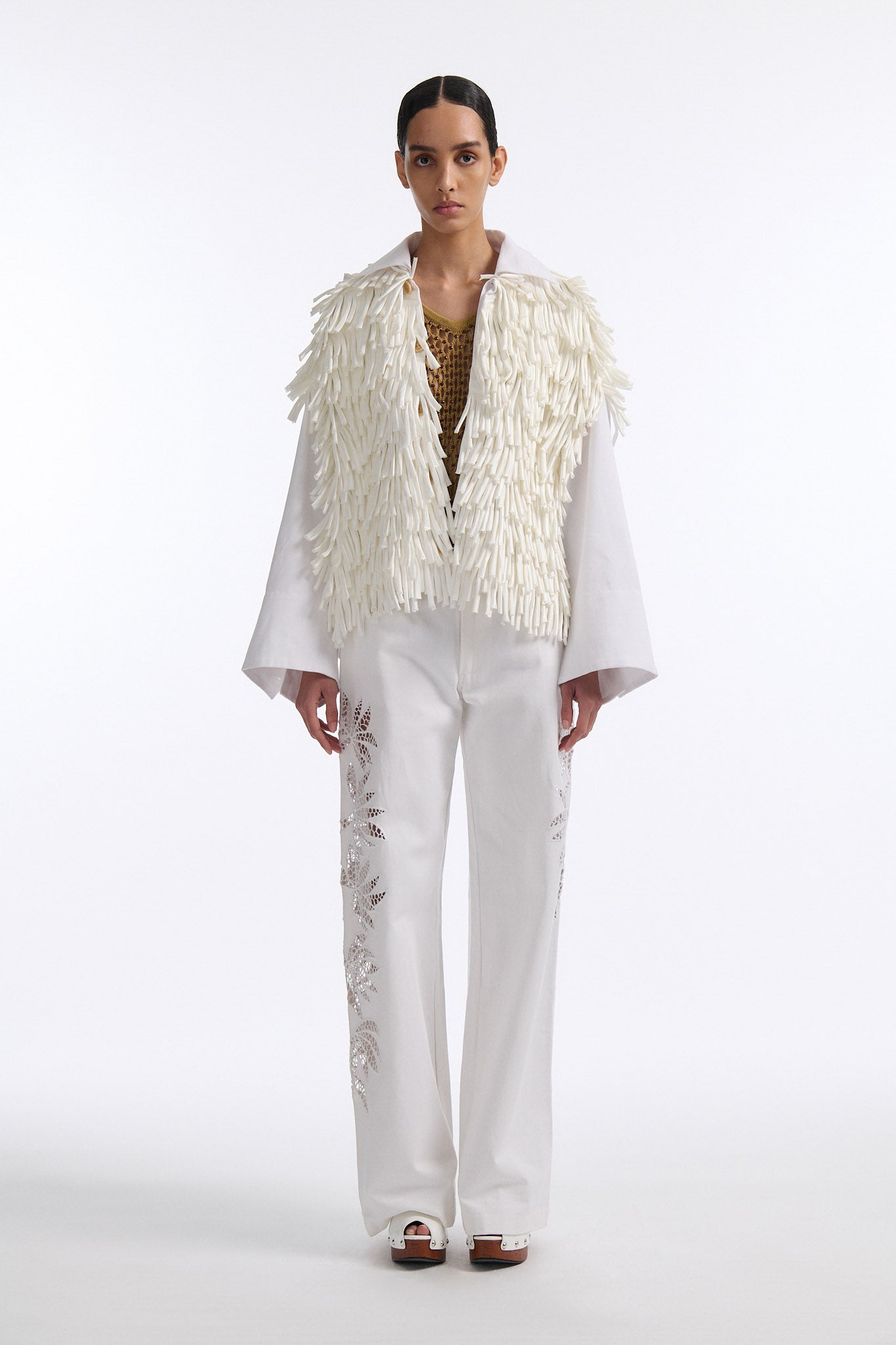 Female model with slicked back black hair in a bun wears white fringe jacket with wide cotton sleeves. The front and back of the jacket are covered in thick white fringe and the sleeves are bare cotton. The model also wears white denim trousers with palm leaf cutouts.
