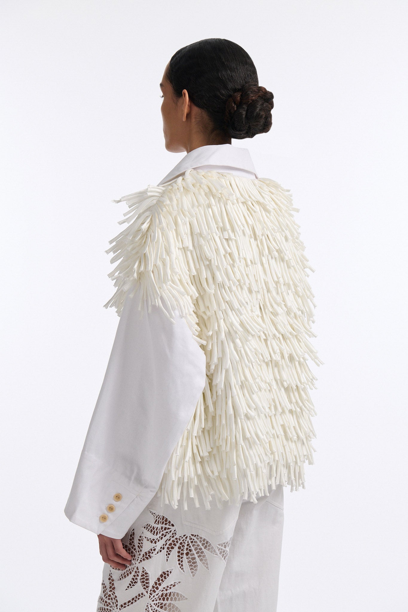 Female model with slicked back black hair in a bun wears white fringe jacket with wide cotton sleeves. The front and back of the jacket are covered in thick white fringe and the sleeves are bare cotton. The model also wears white denim trousers with palm leaf cutouts.