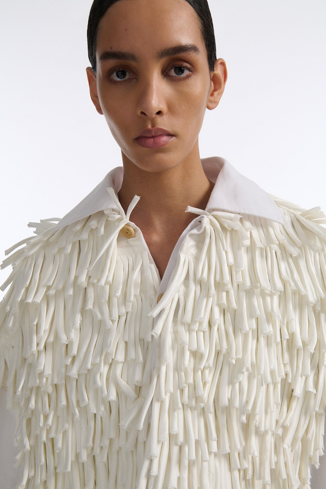 Female model with slicked back black hair in a bun wears white fringe jacket with wide cotton sleeves. The front and back of the jacket are covered in thick white fringe and the sleeves are bare cotton. The model also wears white denim trousers with palm leaf cutouts.