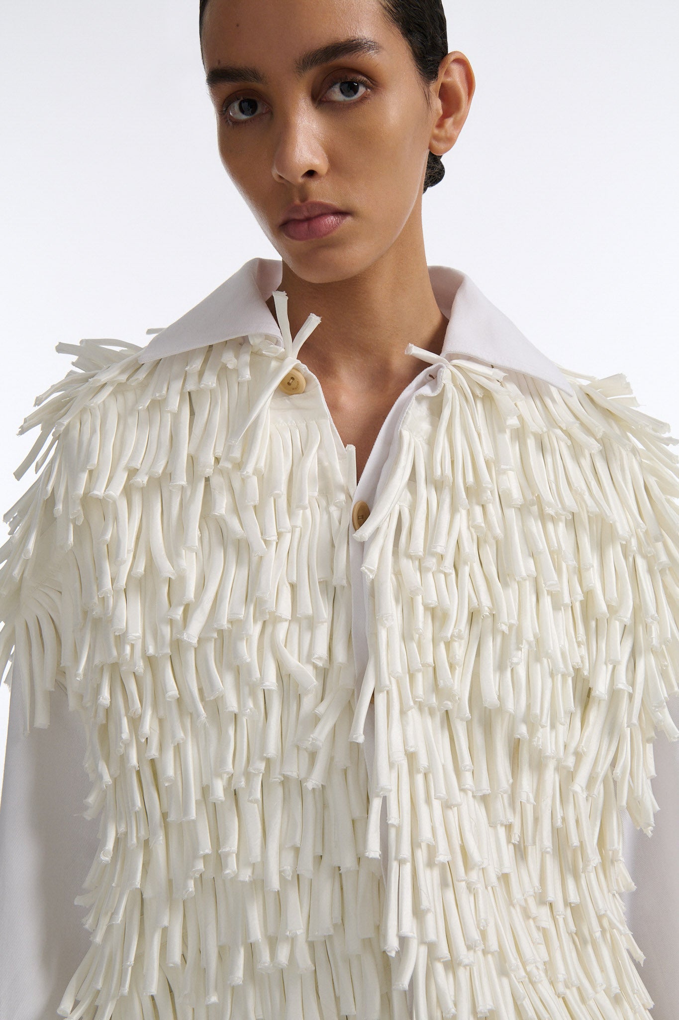 Female model with slicked back black hair in a bun wears white fringe jacket with wide cotton sleeves. The front and back of the jacket are covered in thick white fringe and the sleeves are bare cotton. The model also wears white denim trousers with palm leaf cutouts.