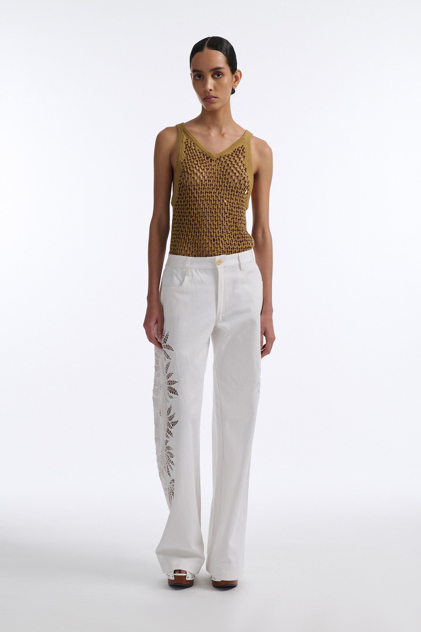 Female model with slicked back black hair in a bun wears cumin colored fine mesh burgundy crystal embellished tank top with palm cutout white denim.