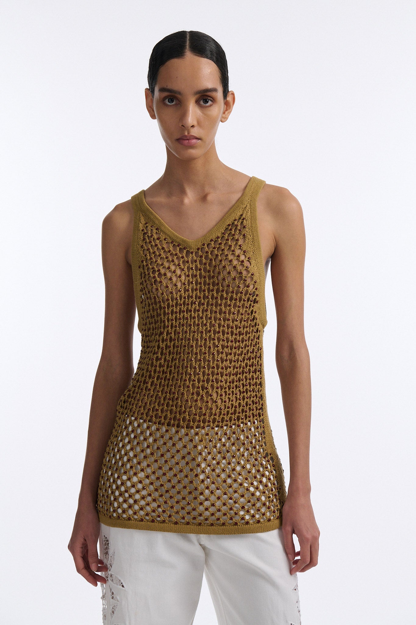 Female model with slicked back black hair in a bun wears cumin colored fine mesh burgundy crystal embellished tank top with palm cutout white denim.