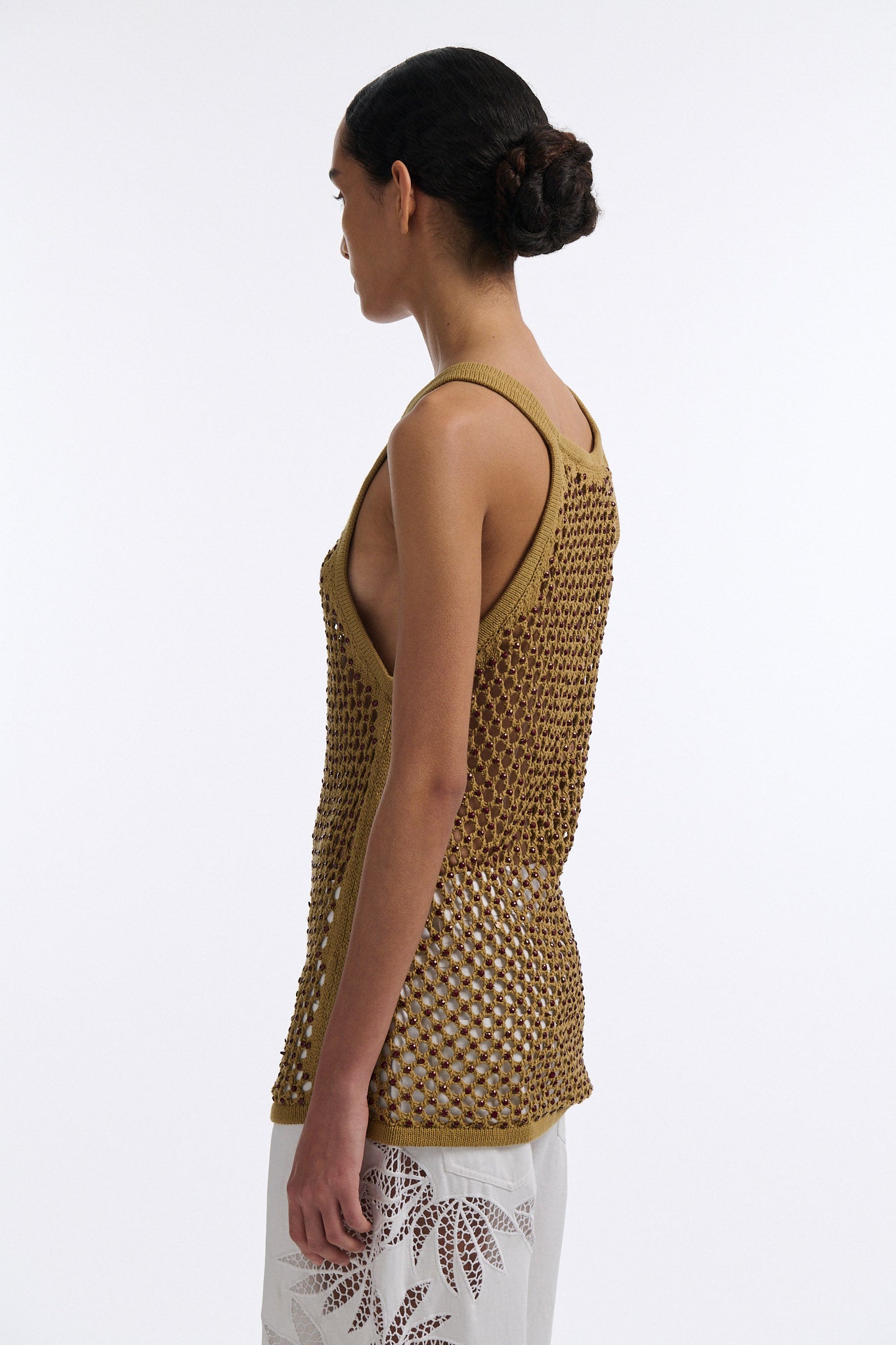 Female model with slicked back black hair in a bun wears cumin colored fine mesh burgundy crystal embellished tank top with palm cutout white denim.