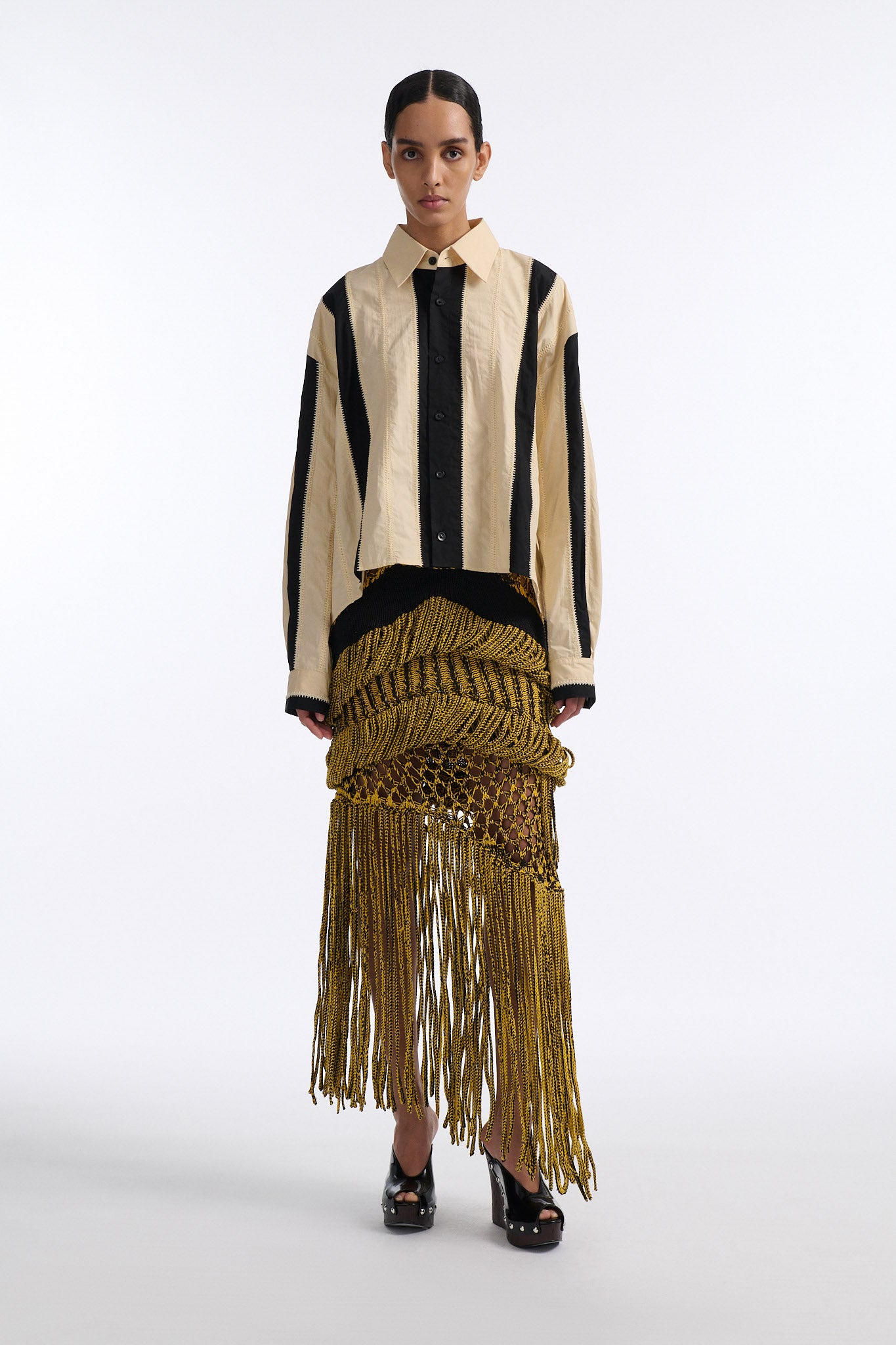 Female model with slicked back black hair in a bun wears ivory and black wide vertical stripe collared shirt. Shirt is wide cut with slight crop. Model also wears mixed stitch mesh skirt made in a yellow and black yarn with long fringe hem.