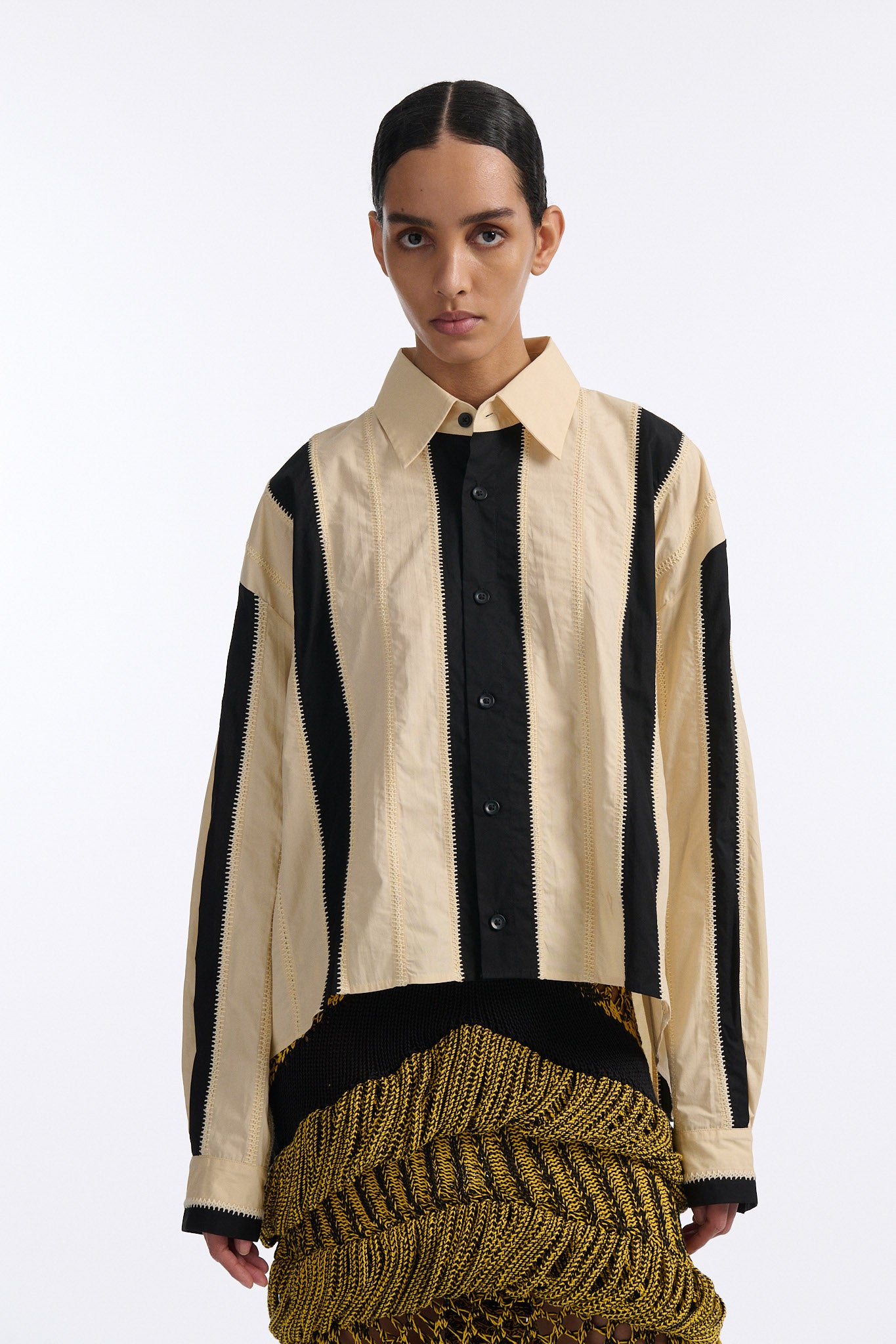 Female model with slicked back black hair in a bun wears ivory and black wide vertical stripe collared shirt. Shirt is wide cut with slight crop. Model also wears mixed stitch mesh skirt made in a yellow and black yarn with long fringe hem.