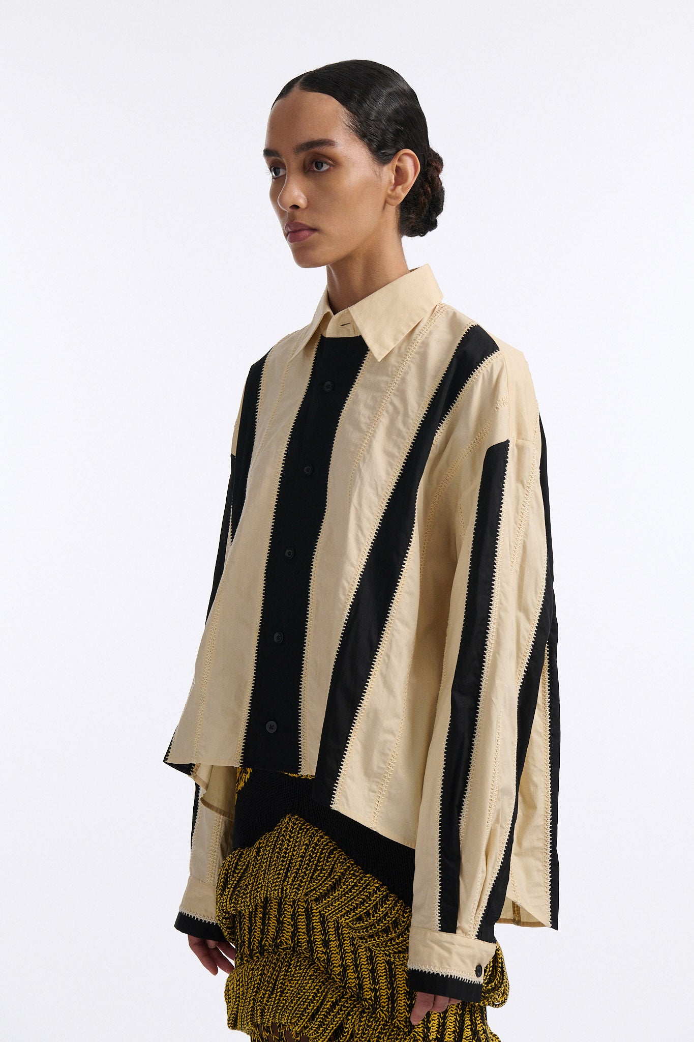 Female model with slicked back black hair in a bun wears ivory and black wide vertical stripe collared shirt. Shirt is wide cut with slight crop. Model also wears mixed stitch mesh skirt made in a yellow and black yarn with long fringe hem.