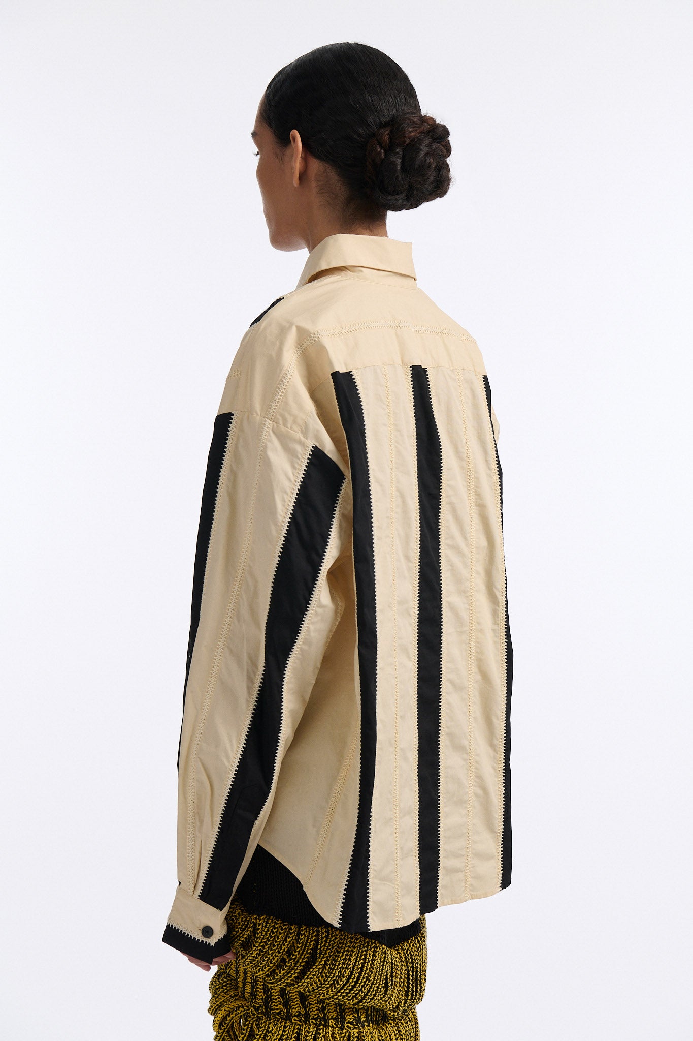 Female model with slicked back black hair in a bun wears ivory and black wide vertical stripe collared shirt. Shirt is wide cut with slight crop. Model also wears mixed stitch mesh skirt made in a yellow and black yarn with long fringe hem.