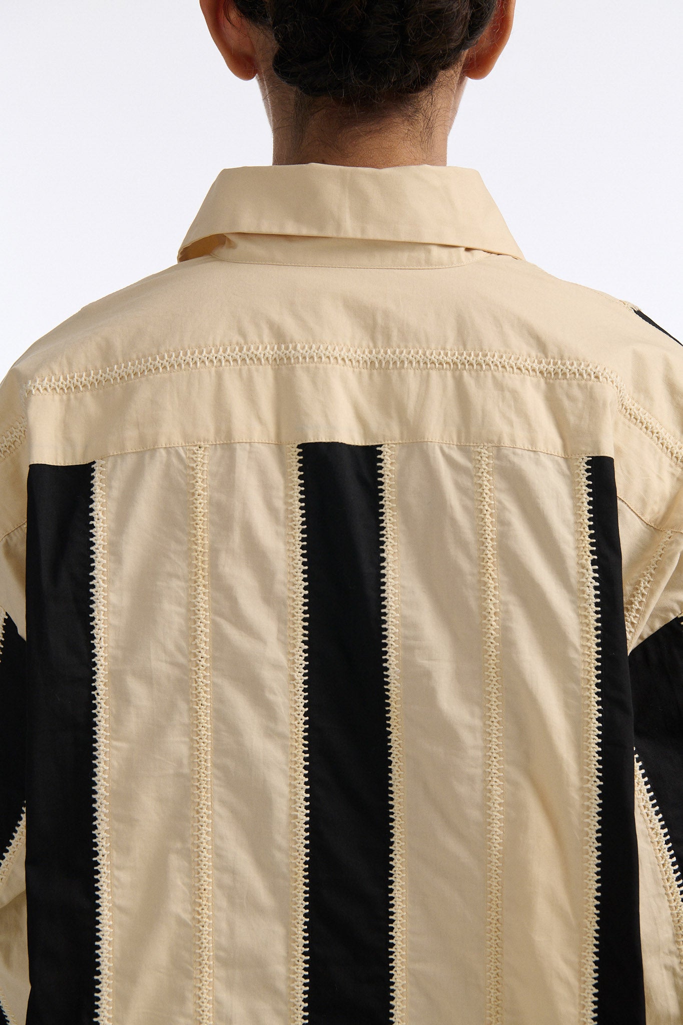 Female model with slicked back black hair in a bun wears ivory and black wide vertical stripe collared shirt. Shirt is wide cut with slight crop. Model also wears mixed stitch mesh skirt made in a yellow and black yarn with long fringe hem.