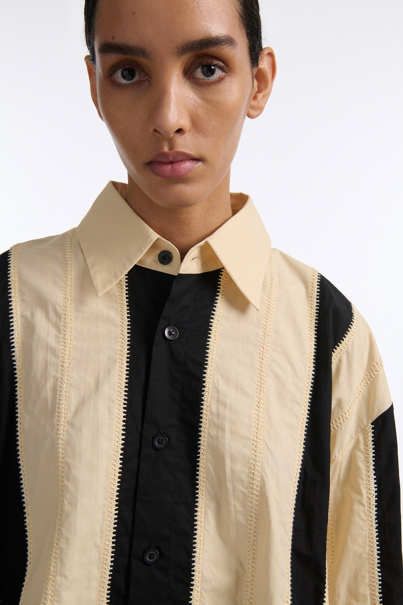 Female model with slicked back black hair in a bun wears ivory and black wide vertical stripe collared shirt. Shirt is wide cut with slight crop. Model also wears mixed stitch mesh skirt made in a yellow and black yarn with long fringe hem.