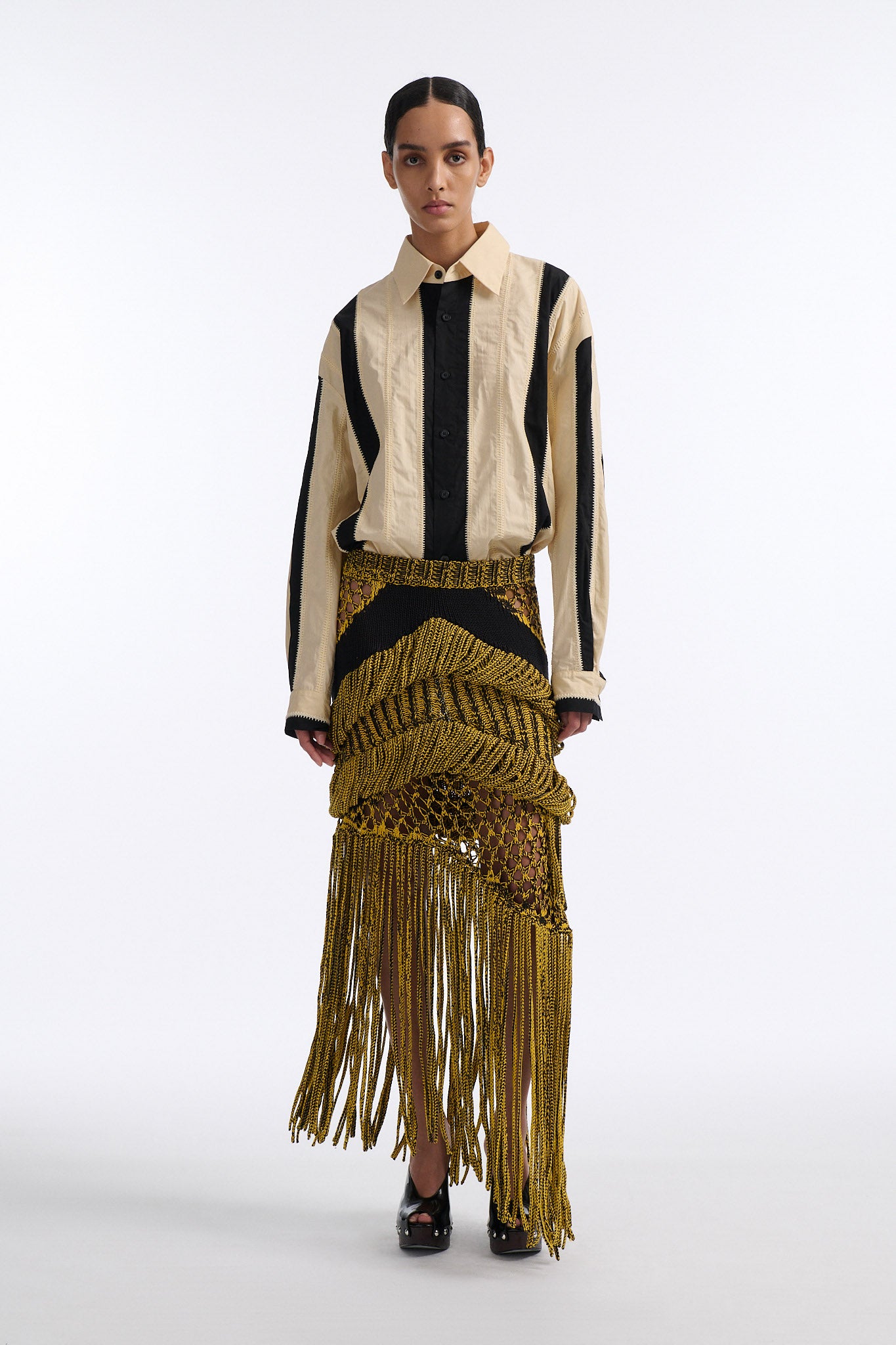 Female model with slicked back black hair in a bun wears ivory and black wide vertical stripe collared shirt. Shirt is wide cut with slight crop. Model also wears mixed stitch mesh skirt made in a yellow and black yarn with long fringe hem.