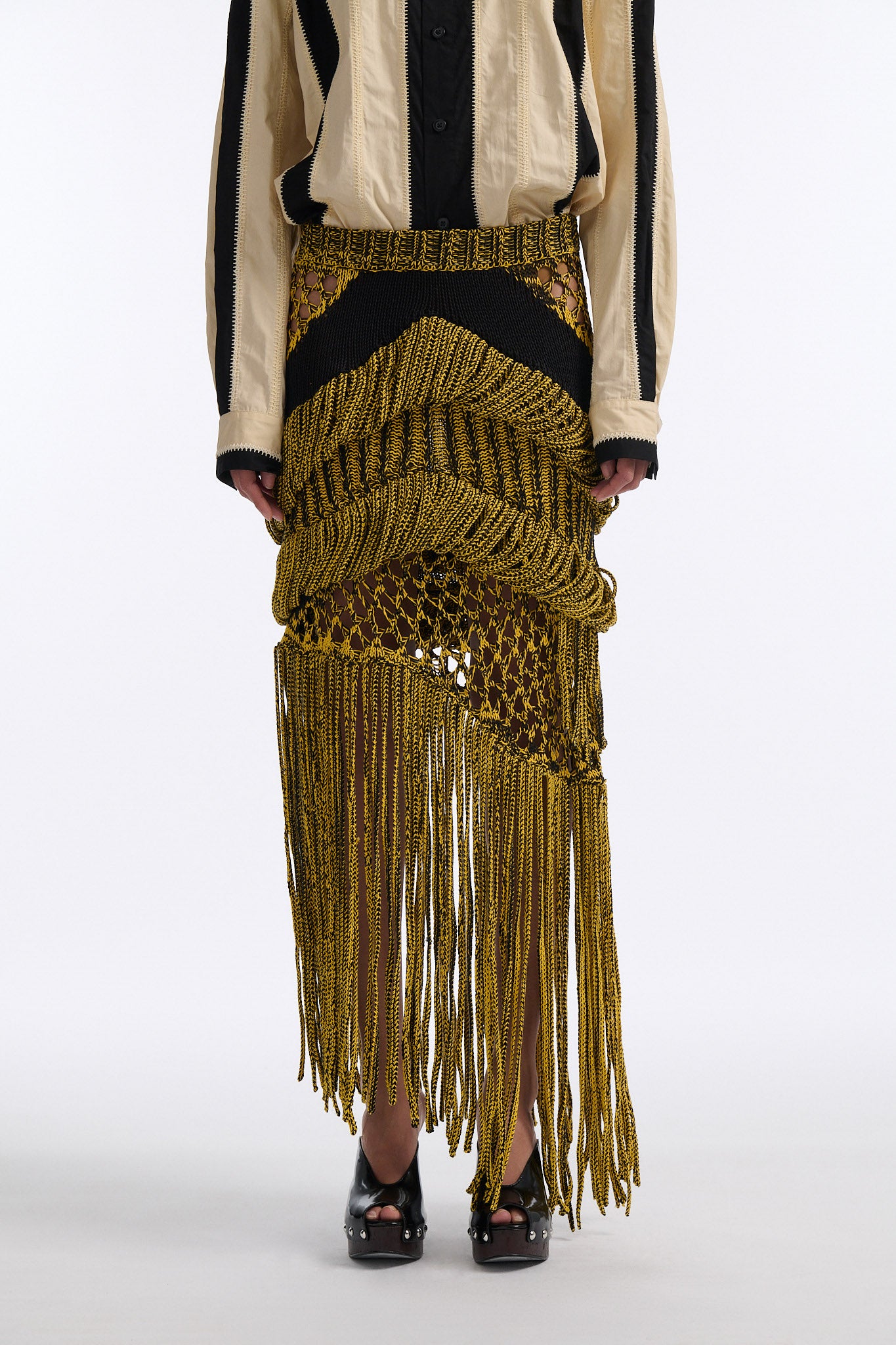 Female model with slicked back black hair in a bun wears ivory and black wide vertical stripe collared shirt. Shirt is wide cut with slight crop. Model also wears mixed stitch mesh skirt made in a yellow and black yarn with long fringe hem.