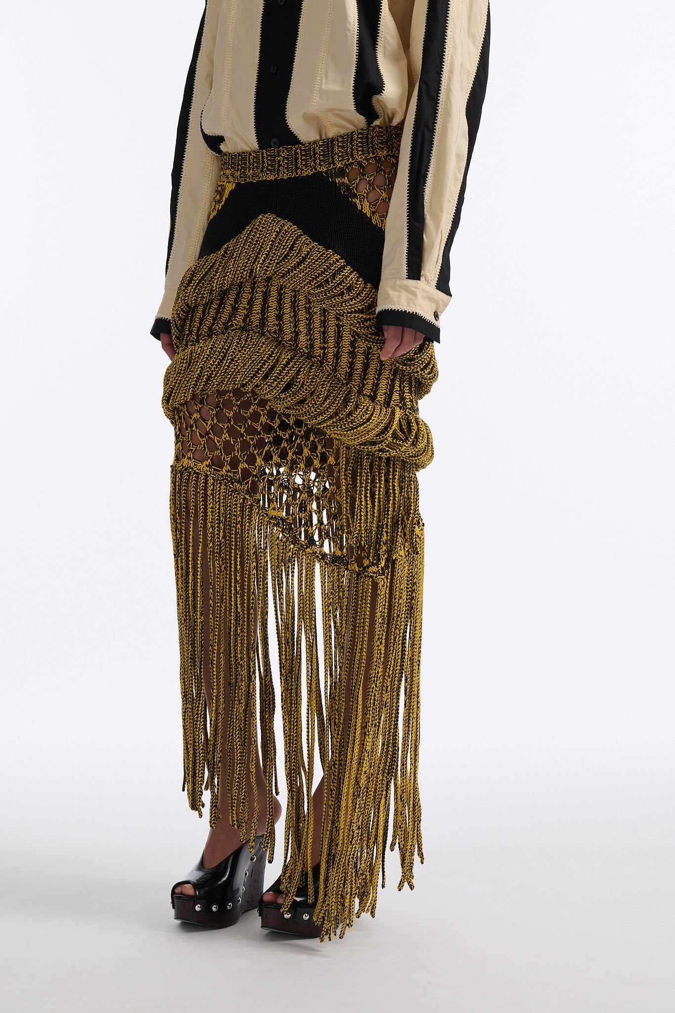 Female model with slicked back black hair in a bun wears ivory and black wide vertical stripe collared shirt. Shirt is wide cut with slight crop. Model also wears mixed stitch mesh skirt made in a yellow and black yarn with long fringe hem.