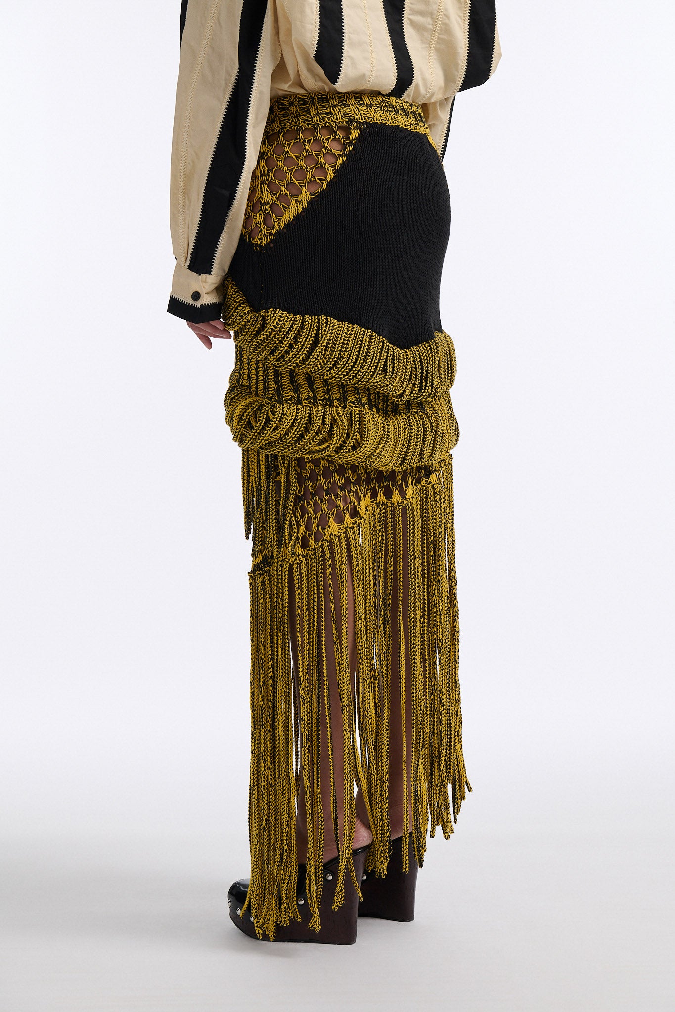 Female model with slicked back black hair in a bun wears ivory and black wide vertical stripe collared shirt. Shirt is wide cut with slight crop. Model also wears mixed stitch mesh skirt made in a yellow and black yarn with long fringe hem.