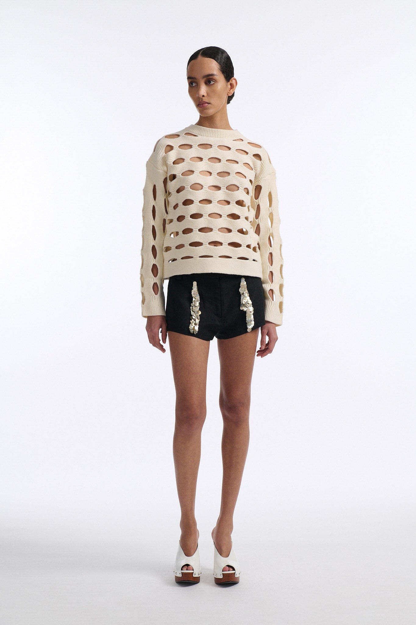 Female model with slicked back black hair in a bun wears black linen shorts with vertical shell embellishments on the front. The shorts have a 2 inch inseam and are worn with an allover cutout off-white sweater.