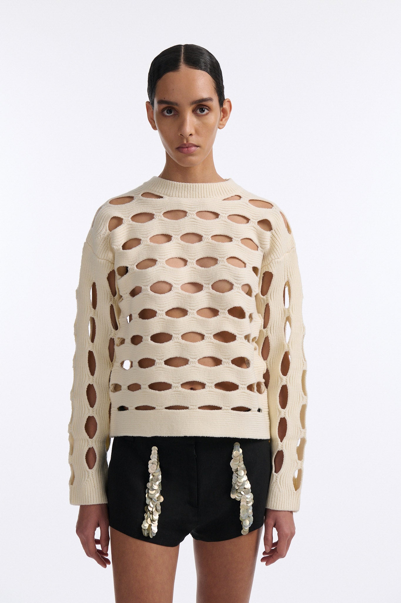 Female model with slicked back black hair in a bun wears black linen shorts with vertical shell embellishments on the front. The shorts have a 2 inch inseam and are worn with an allover cutout off-white sweater.