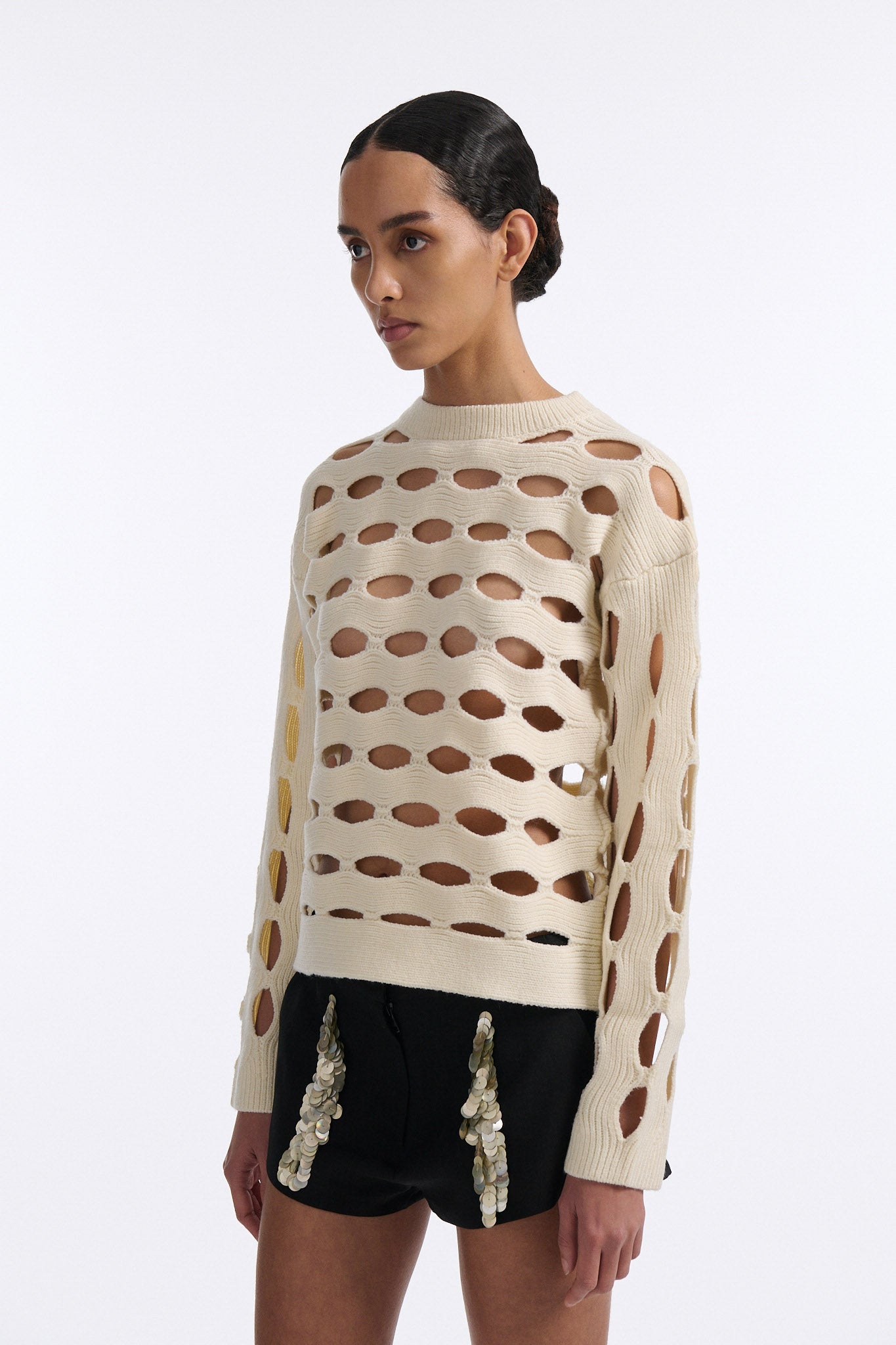 Female model with slicked back black hair in a bun wears black linen shorts with vertical shell embellishments on the front. The shorts have a 2 inch inseam and are worn with an allover cutout off-white sweater.