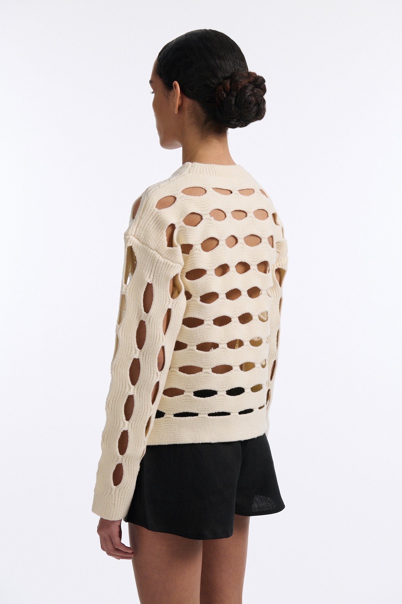 Female model with slicked back black hair in a bun wears black linen shorts with vertical shell embellishments on the front. The shorts have a 2 inch inseam and are worn with an allover cutout off-white sweater.