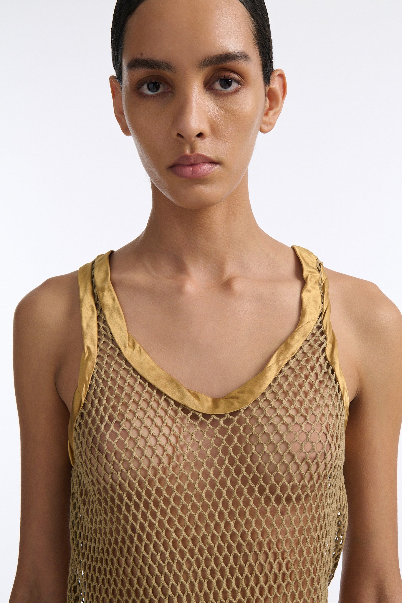 Female model with slicked back black hair in a bun wears golden colored cotton mesh see-through top with wide silk binding at hems. Model also wears white crochet skirt and tall wedges.
