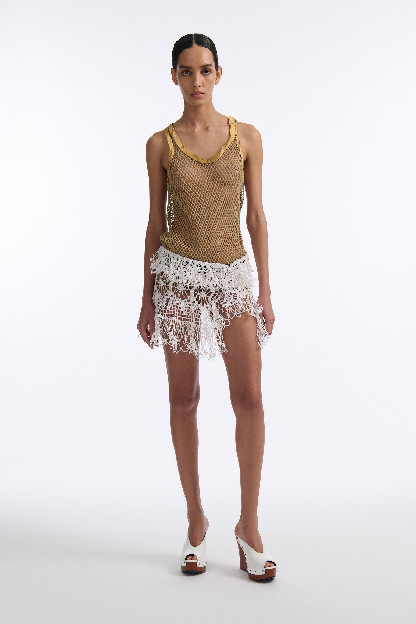 Female model with slicked back black hair in a bun wears white crochet see-through mini skirt and gold mesh tank top with silk detailing.