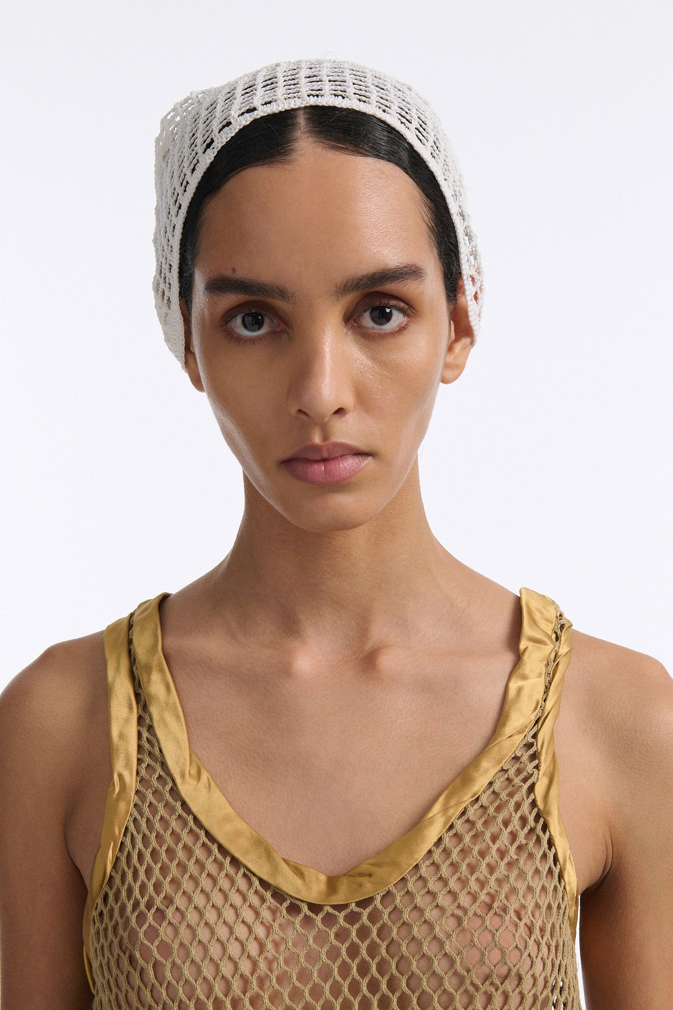 Female model with slicked back black hair in a bun wears gold see-through mesh tank top and white crochet bandana tied around her forehead.