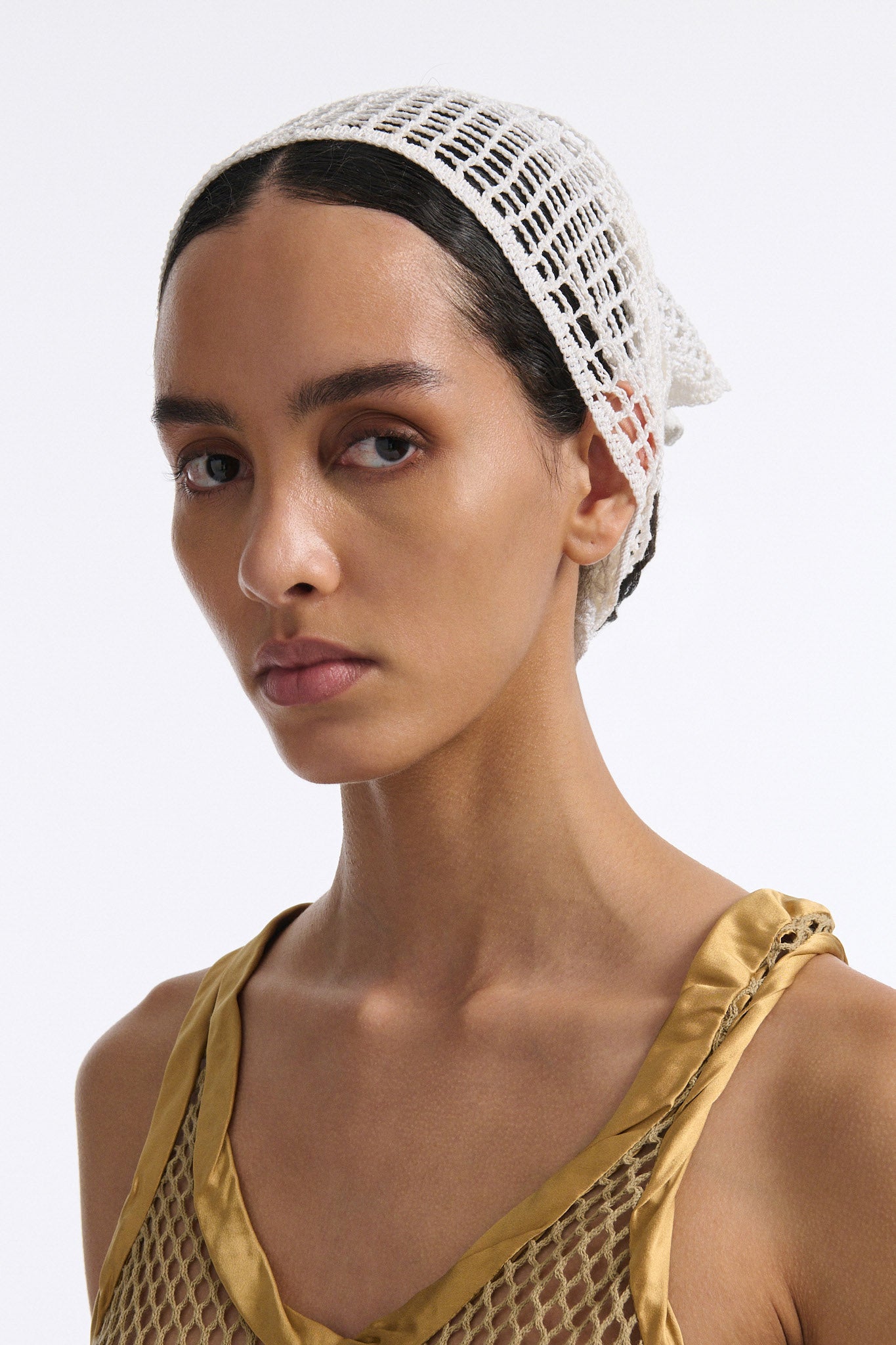 Female model with slicked back black hair in a bun wears gold see-through mesh tank top and white crochet bandana tied around her forehead.