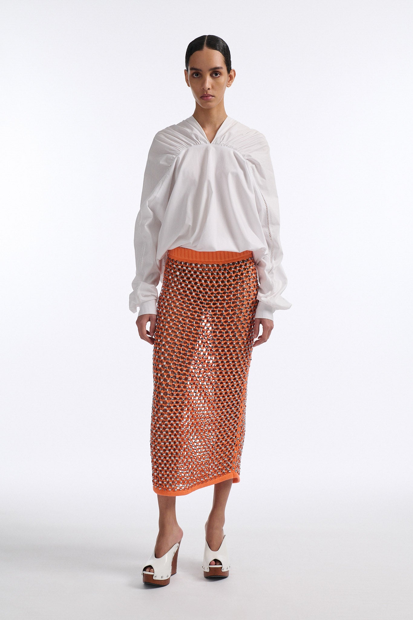 Female model with slicked back black hair in a bun wears white gathered V-neck blouse with puff sleeves and drawstring at waist. Model also wears bright orange chunky mesh skirt with clear crystal hand-sewn embellishments.