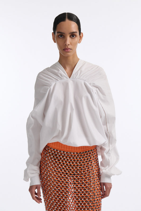 Female model with slicked back black hair in a bun wears white gathered V-neck blouse with puff sleeves and drawstring at waist. Model also wears bright orange chunky mesh skirt with clear crystal hand-sewn embellishments.