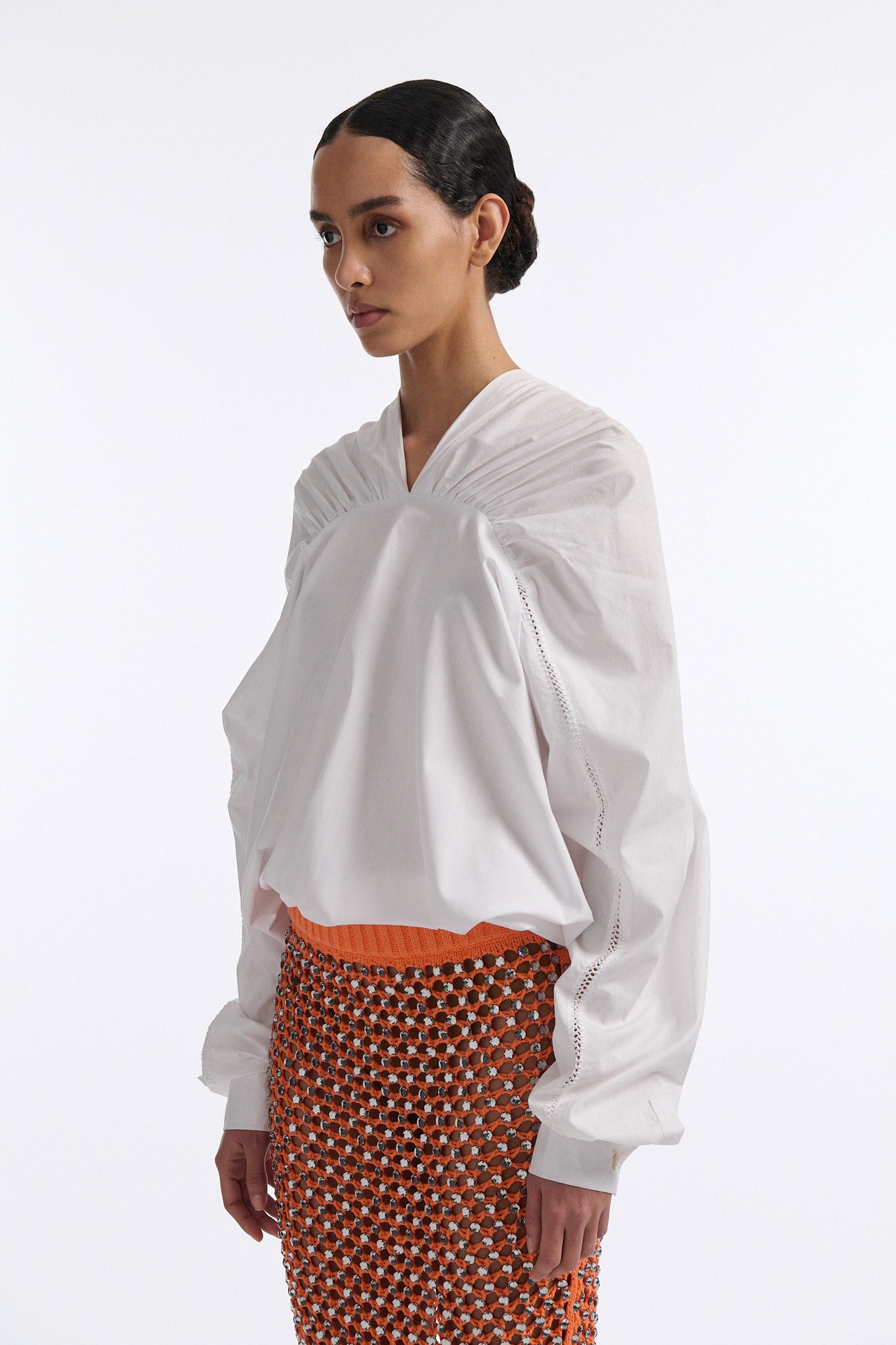 Female model with slicked back black hair in a bun wears white gathered V-neck blouse with puff sleeves and drawstring at waist. Model also wears bright orange chunky mesh skirt with clear crystal hand-sewn embellishments.