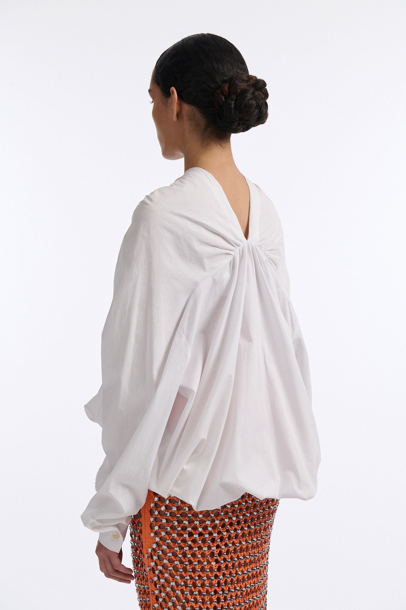 Female model with slicked back black hair in a bun wears white gathered V-neck blouse with puff sleeves and drawstring at waist. Model also wears bright orange chunky mesh skirt with clear crystal hand-sewn embellishments.