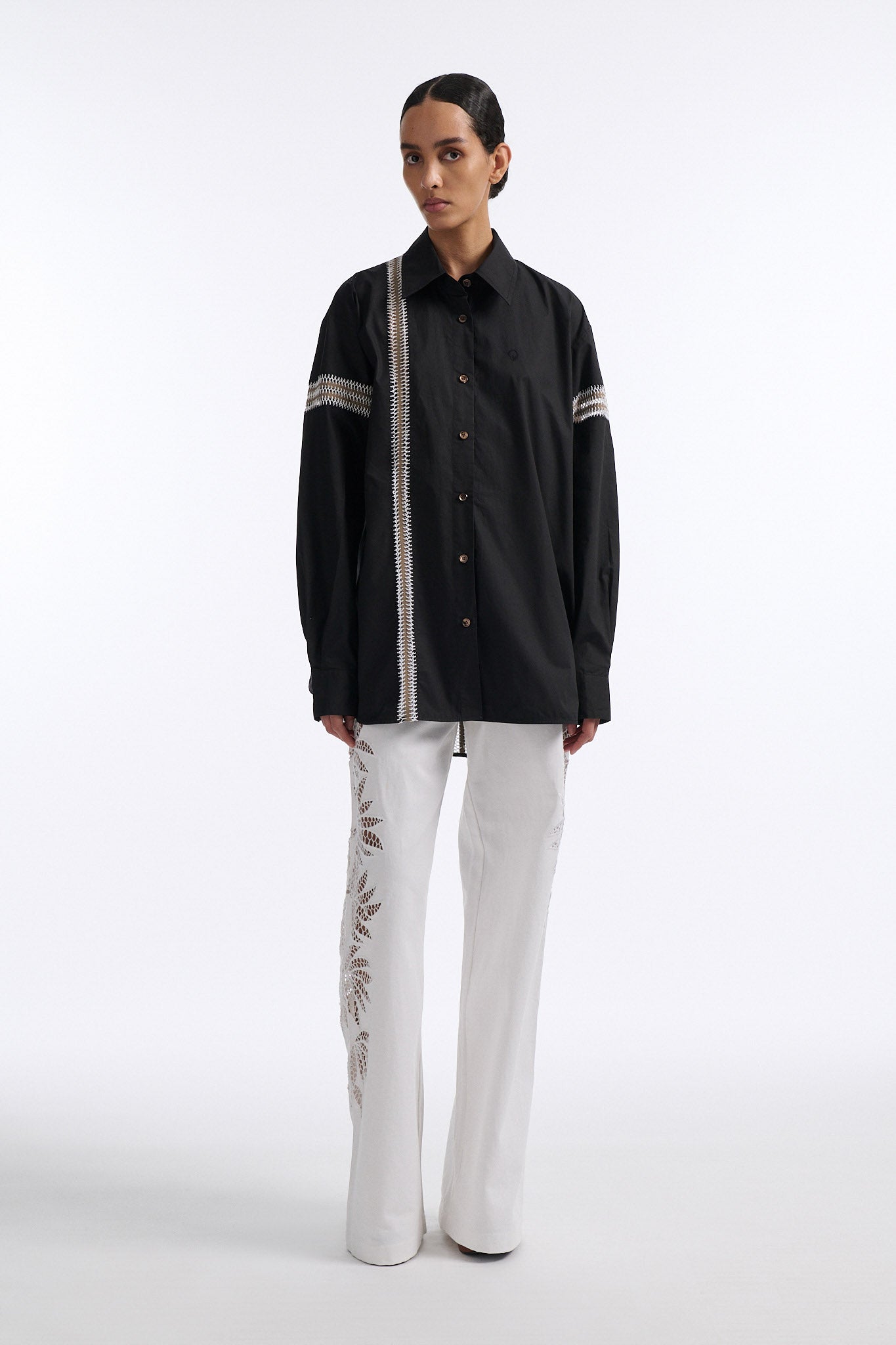 Female model with slicked back black hair in a bun wears black long sleeve button down shirt with white hand stitched detailing along left and on the tops of both sleeves. Model also wears white denim trousers with palm leaf cutouts along both legs.
