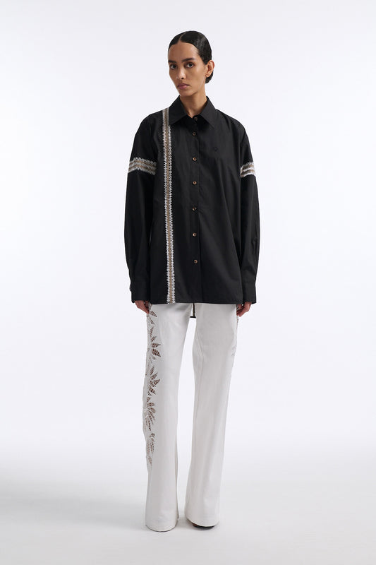 Female model with slicked back black hair in a bun wears black long sleeve button down shirt with white hand stitched detailing along left and on the tops of both sleeves. Model also wears white denim trousers with palm leaf cutouts along both legs.