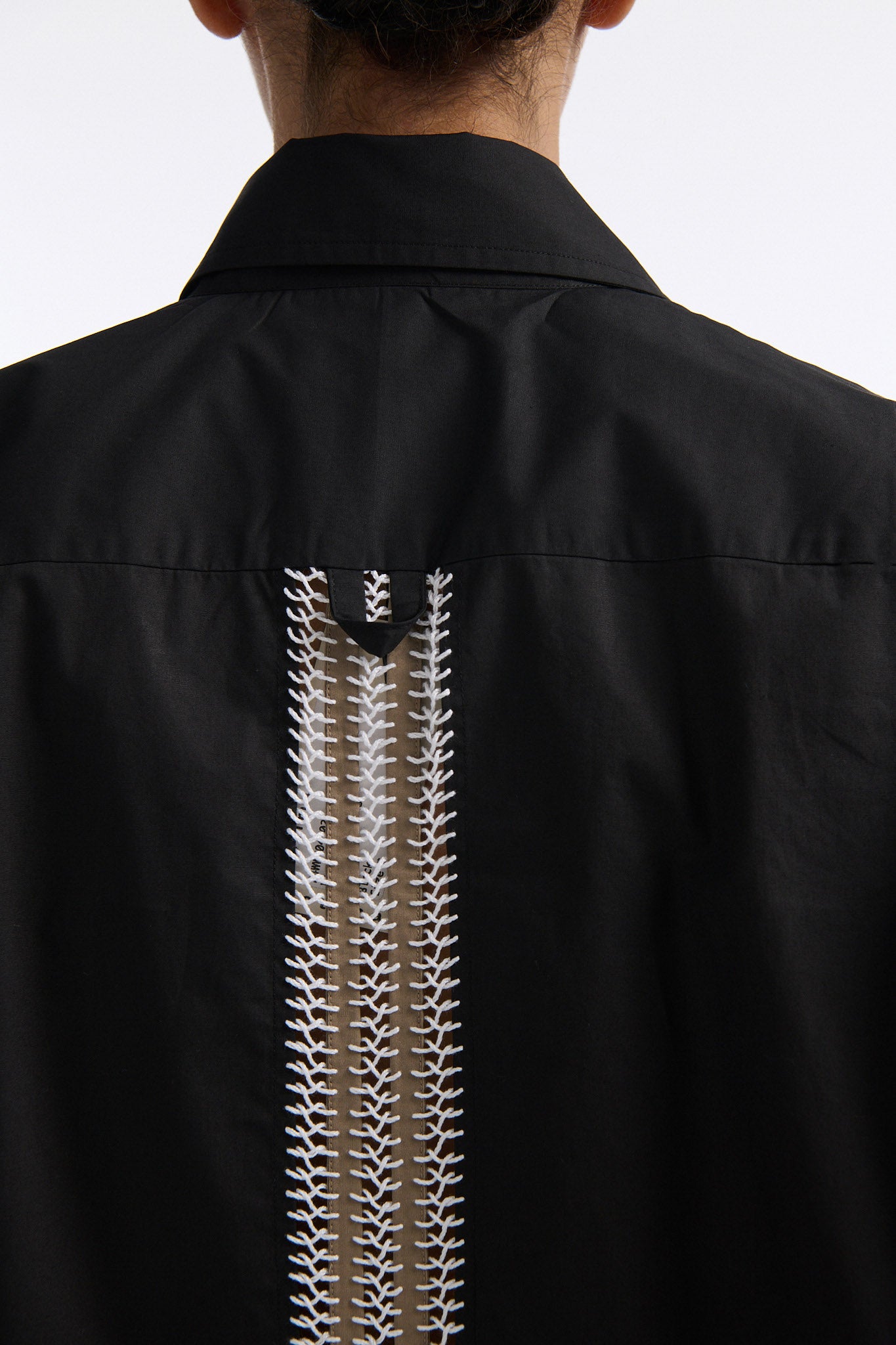 Female model with slicked back black hair in a bun wears black long sleeve button down shirt with white hand stitched detailing along left and on the tops of both sleeves. Model also wears white denim trousers with palm leaf cutouts along both legs.