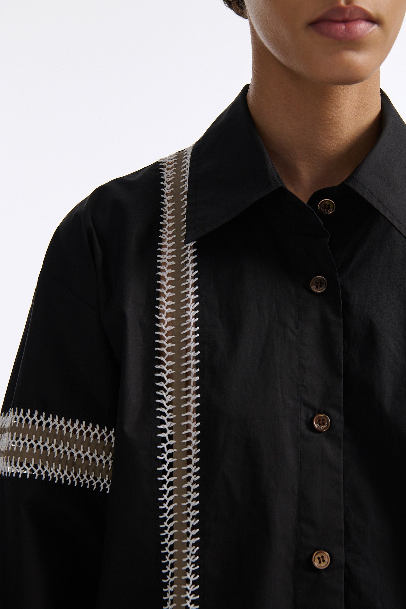 Female model with slicked back black hair in a bun wears black long sleeve button down shirt with white hand stitched detailing along left and on the tops of both sleeves. Model also wears white denim trousers with palm leaf cutouts along both legs.