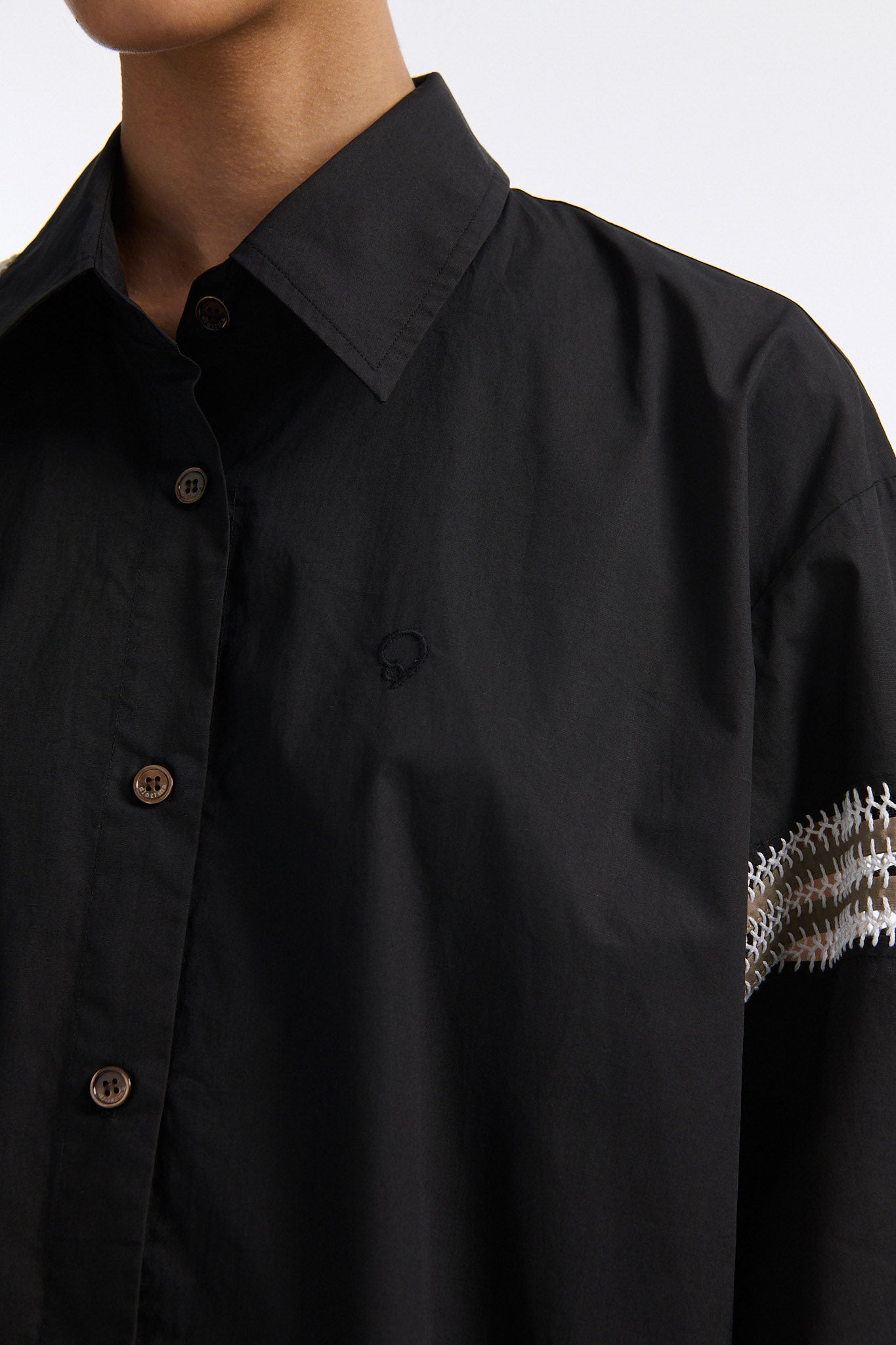 Female model with slicked back black hair in a bun wears black long sleeve button down shirt with white hand stitched detailing along left and on the tops of both sleeves. Model also wears white denim trousers with palm leaf cutouts along both legs.