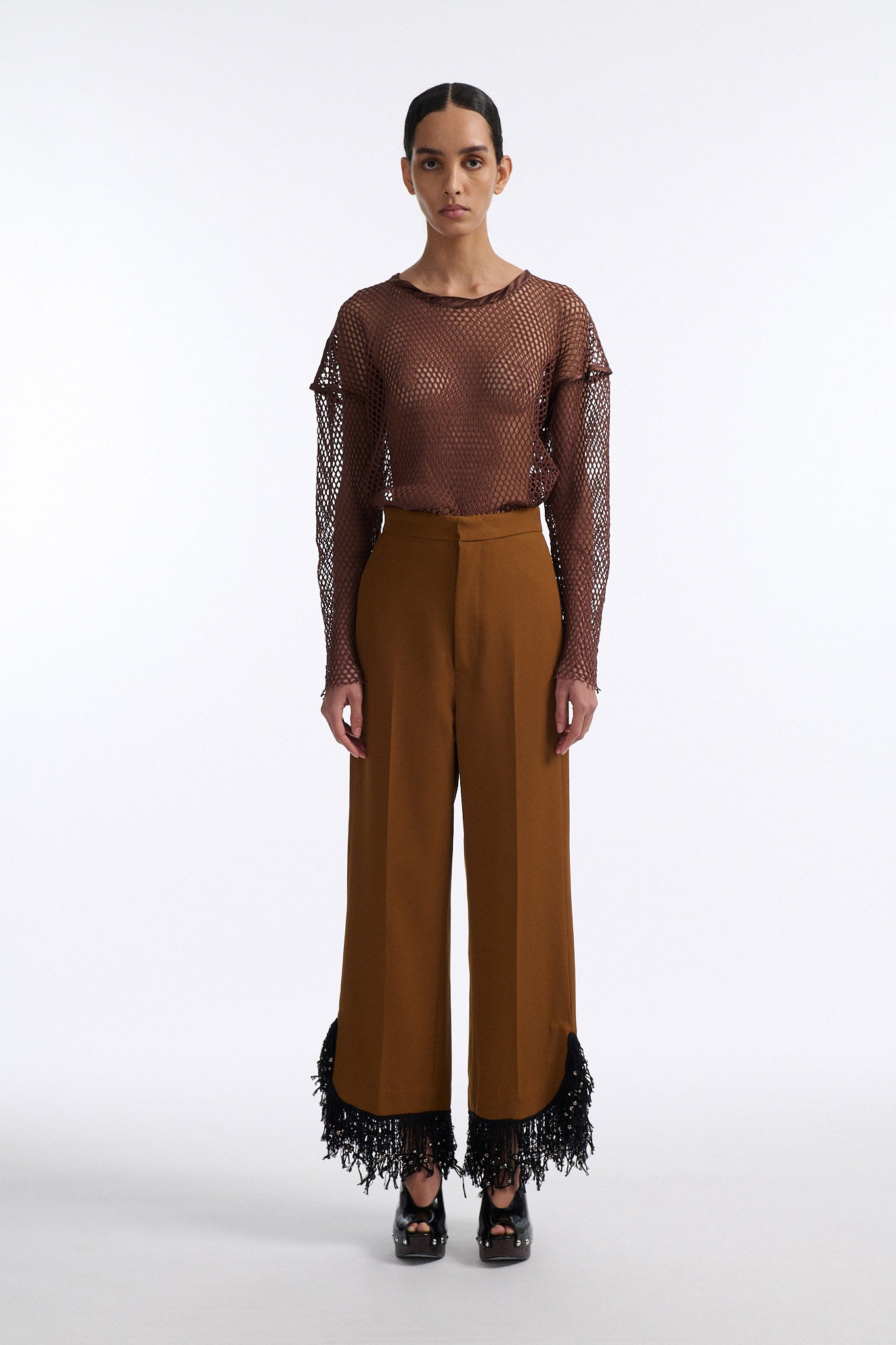 Female model with slicked back black hair in a bun wears brown cotton mesh see-through long sleeve top with silk piping. Model also wears brown tobacco colored high-waisted fine wool trouser with curved hem that ends with beaded black fringe.