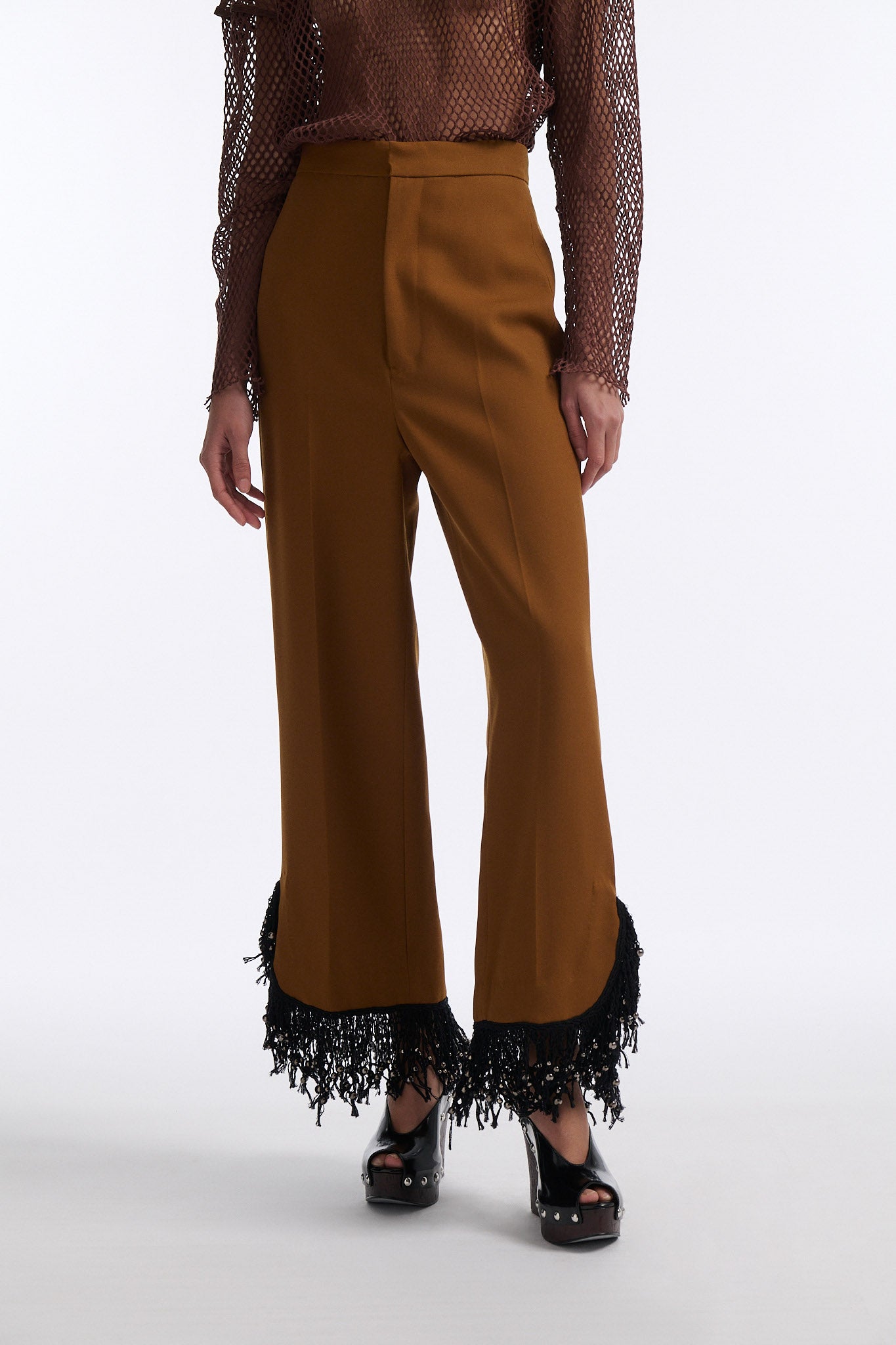 Female model with slicked back black hair in a bun wears brown cotton mesh see-through long sleeve top with silk piping. Model also wears brown tobacco colored high-waisted fine wool trouser with curved hem that ends with beaded black fringe.