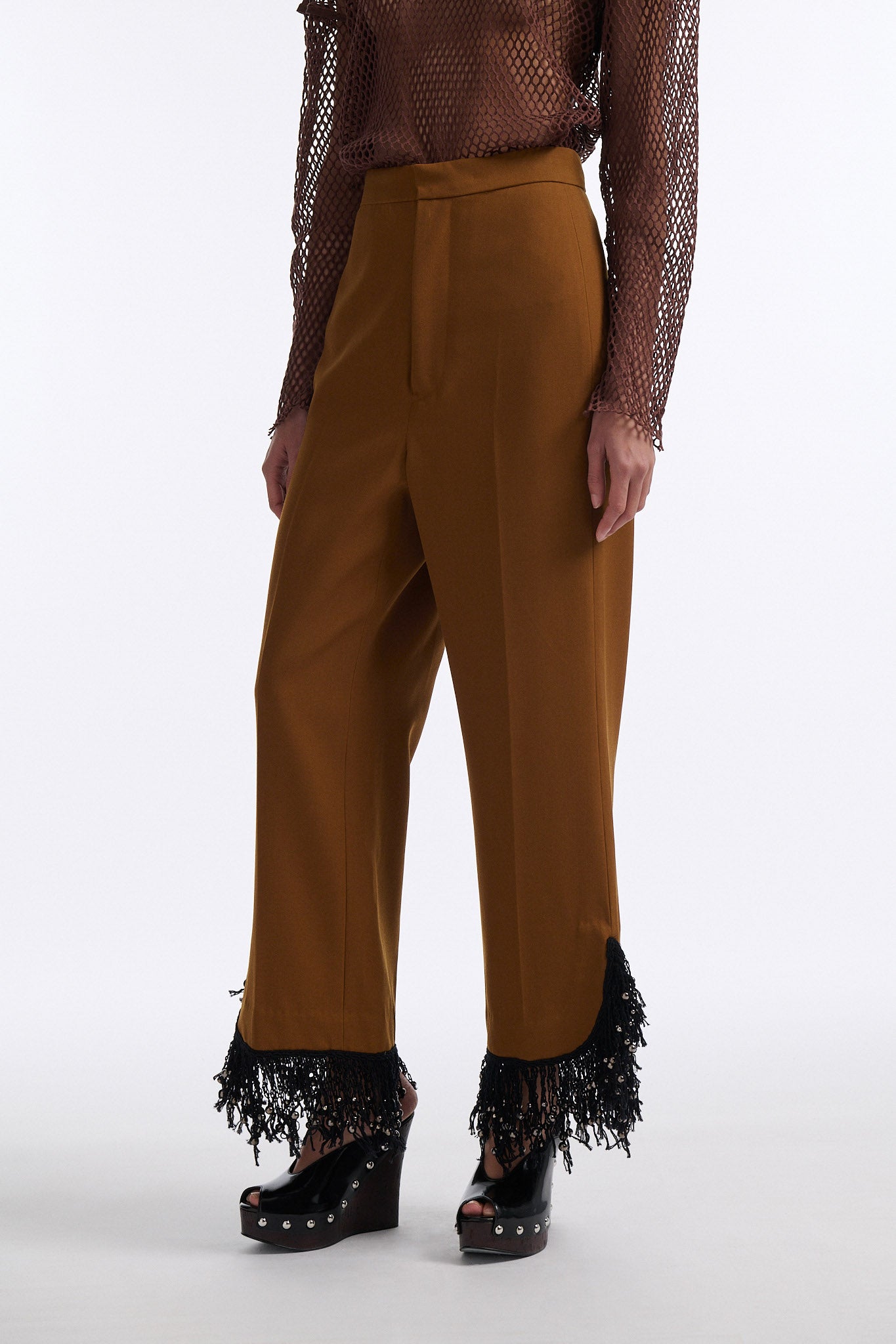 Female model with slicked back black hair in a bun wears brown cotton mesh see-through long sleeve top with silk piping. Model also wears brown tobacco colored high-waisted fine wool trouser with curved hem that ends with beaded black fringe.