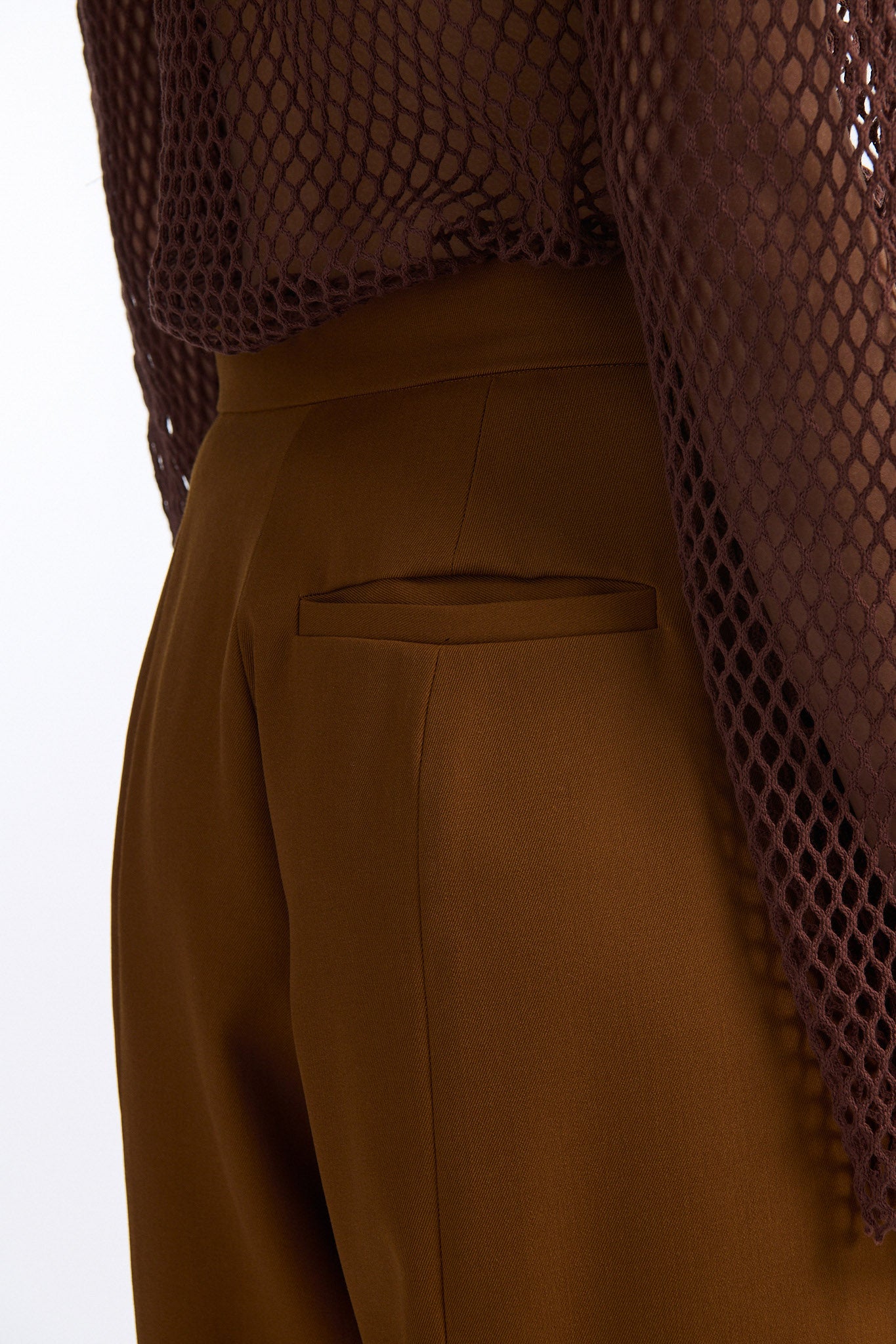 Female model with slicked back black hair in a bun wears brown cotton mesh see-through long sleeve top with silk piping. Model also wears brown tobacco colored high-waisted fine wool trouser with curved hem that ends with beaded black fringe.