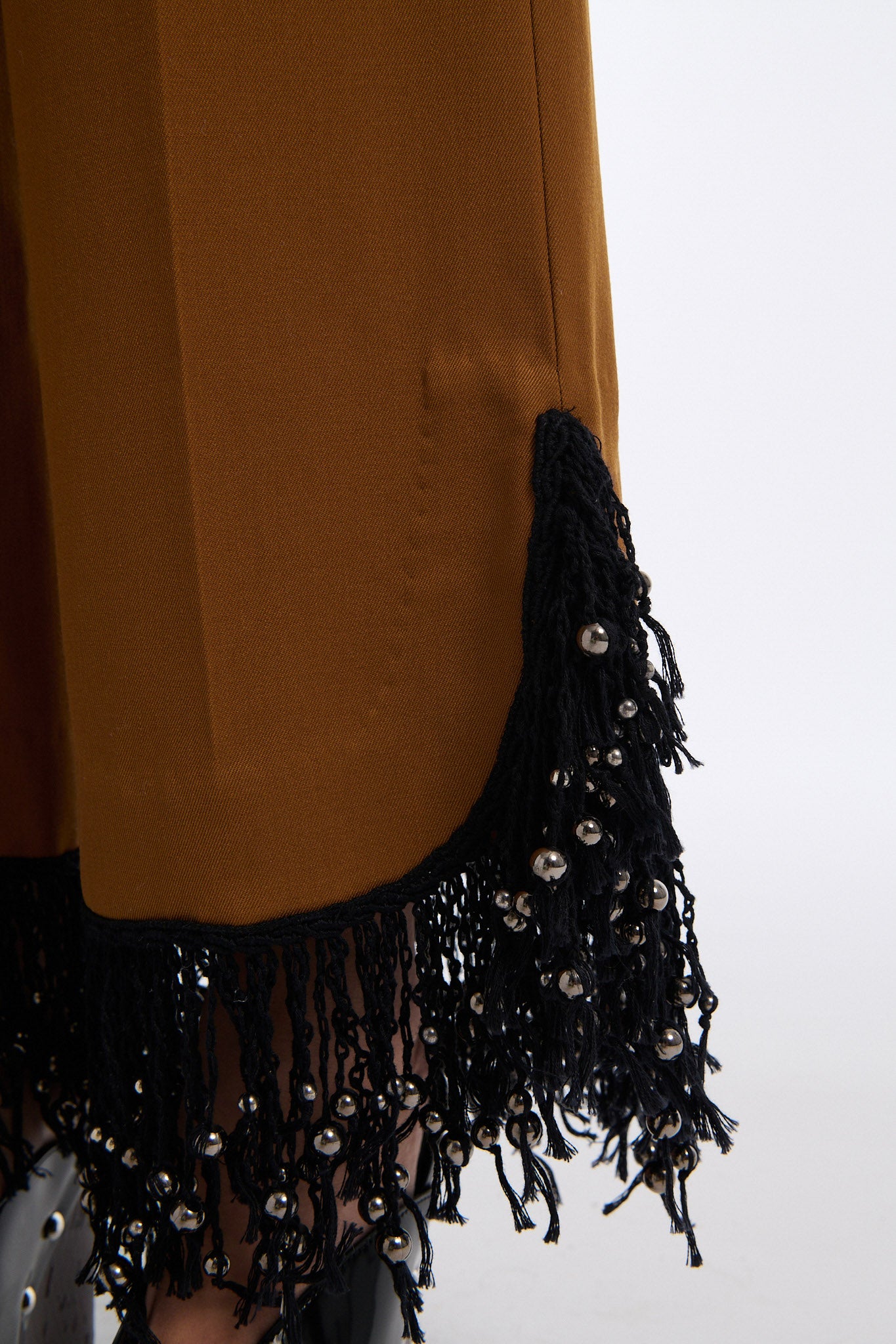 Female model with slicked back black hair in a bun wears brown cotton mesh see-through long sleeve top with silk piping. Model also wears brown tobacco colored high-waisted fine wool trouser with curved hem that ends with beaded black fringe.