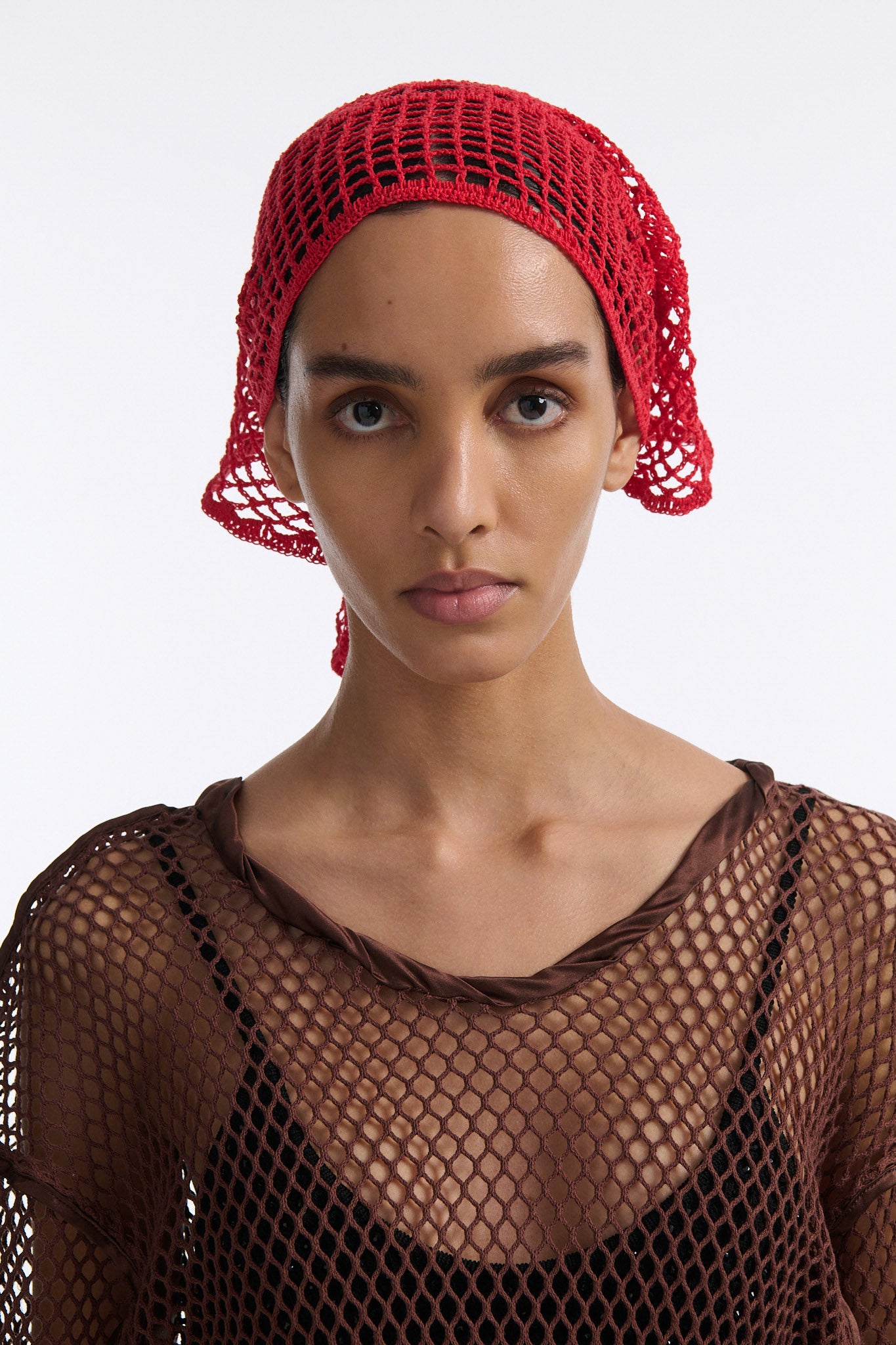Female model with slicked back black hair in a bun wears brown see-through mesh long sleeve top and bright red crochet bandana tied around her forehead.